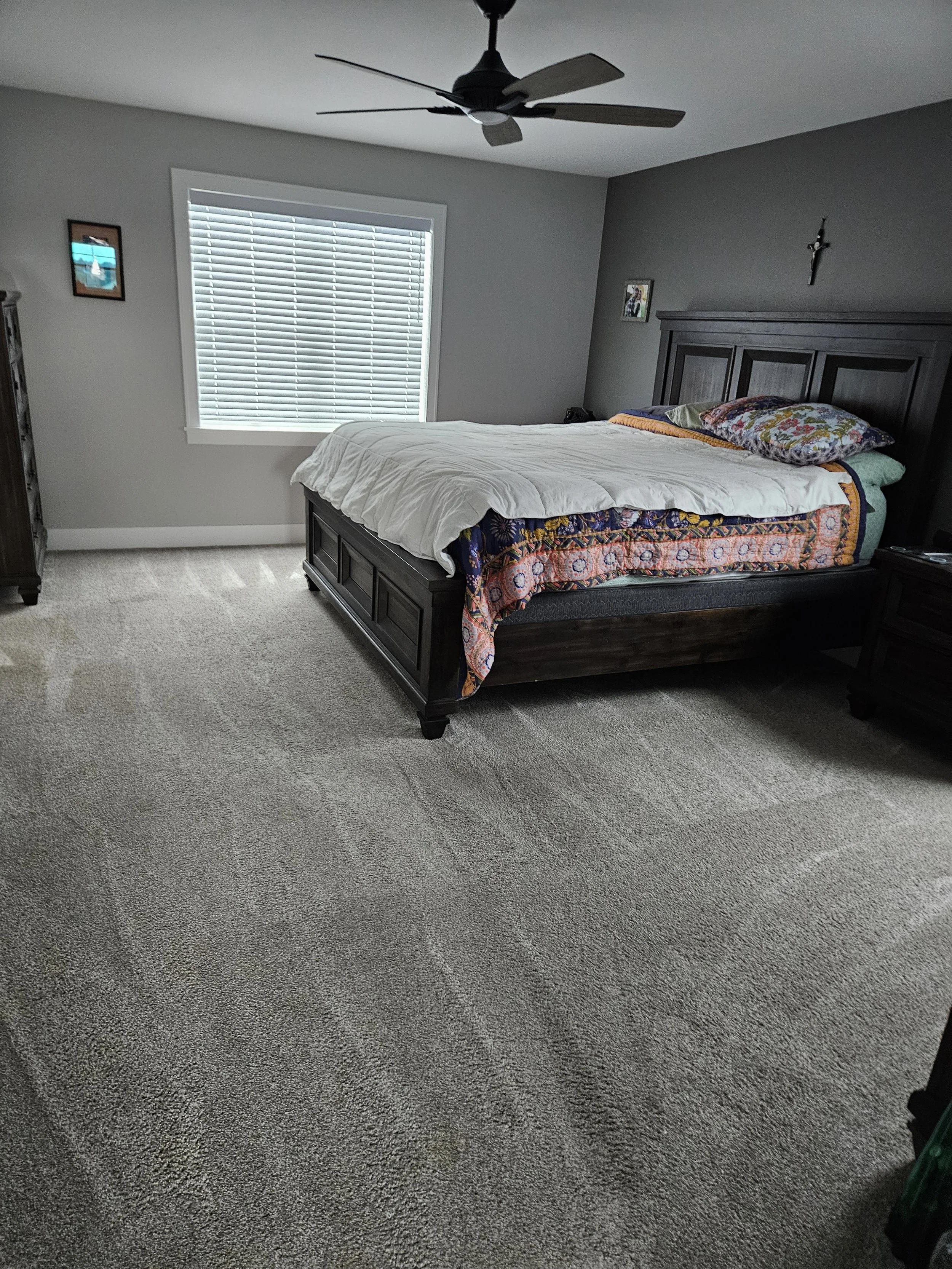 A bedroom with a dark wooden bed frame, dressed with a colorful quilt and white comforter, and one pillow. There is a window with blinds, a dark wooden nightstand, and a ceiling fan. The walls are painted gray, decorated with a crucifix and framed photographs.