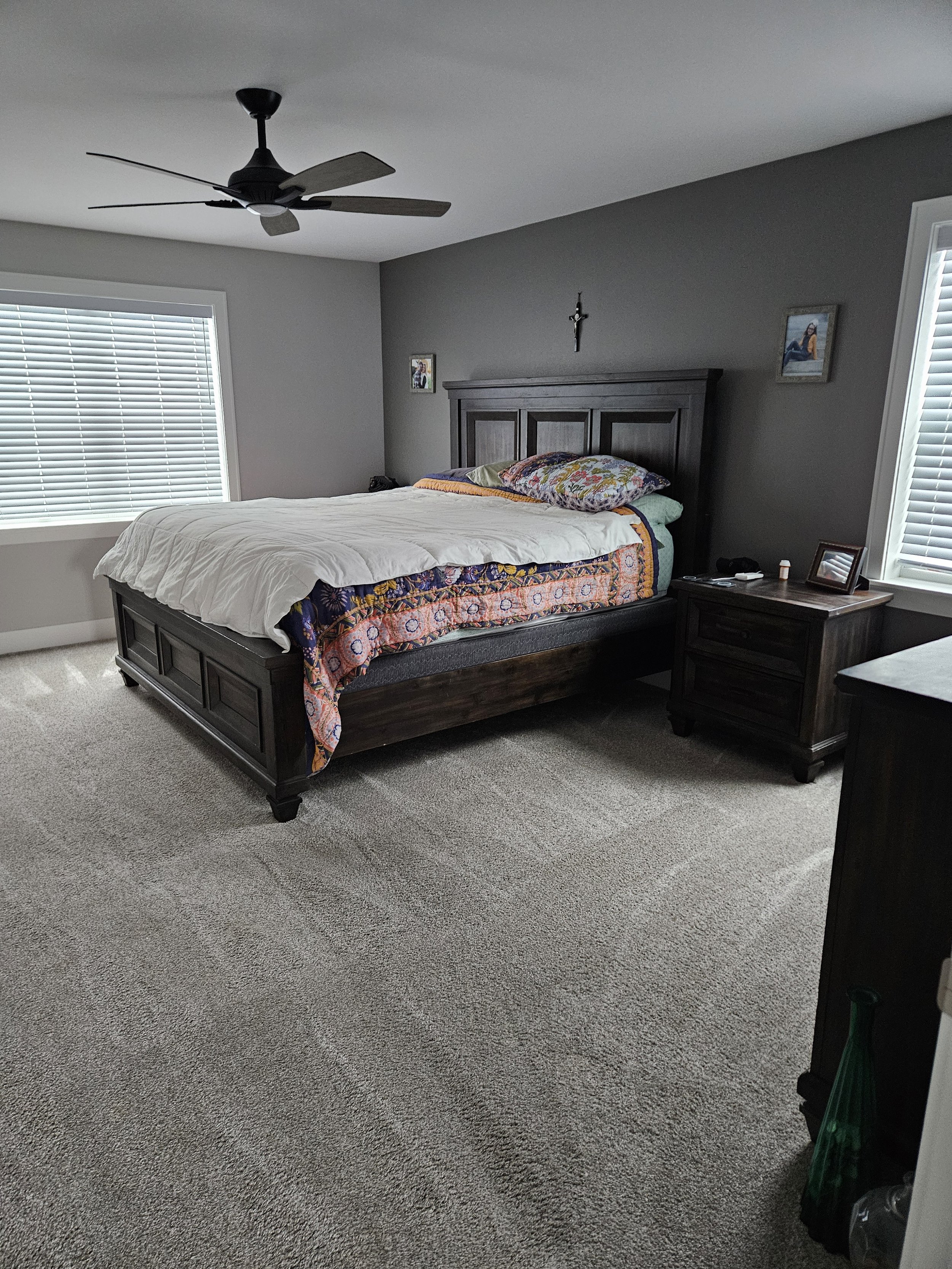 A neatly made bed with a dark wooden frame and headboard in a bedroom with gray walls. There is a colorful quilt, a white comforter, and pillows on the bed. A small dark wooden nightstand with a framed photo, remote, and small bottles sits beside the bed. Two windows with blinds are on either side, letting in natural light. A ceiling fan with dark blades is on the ceiling, and there are small pictures and a cross on the wall above the bed.