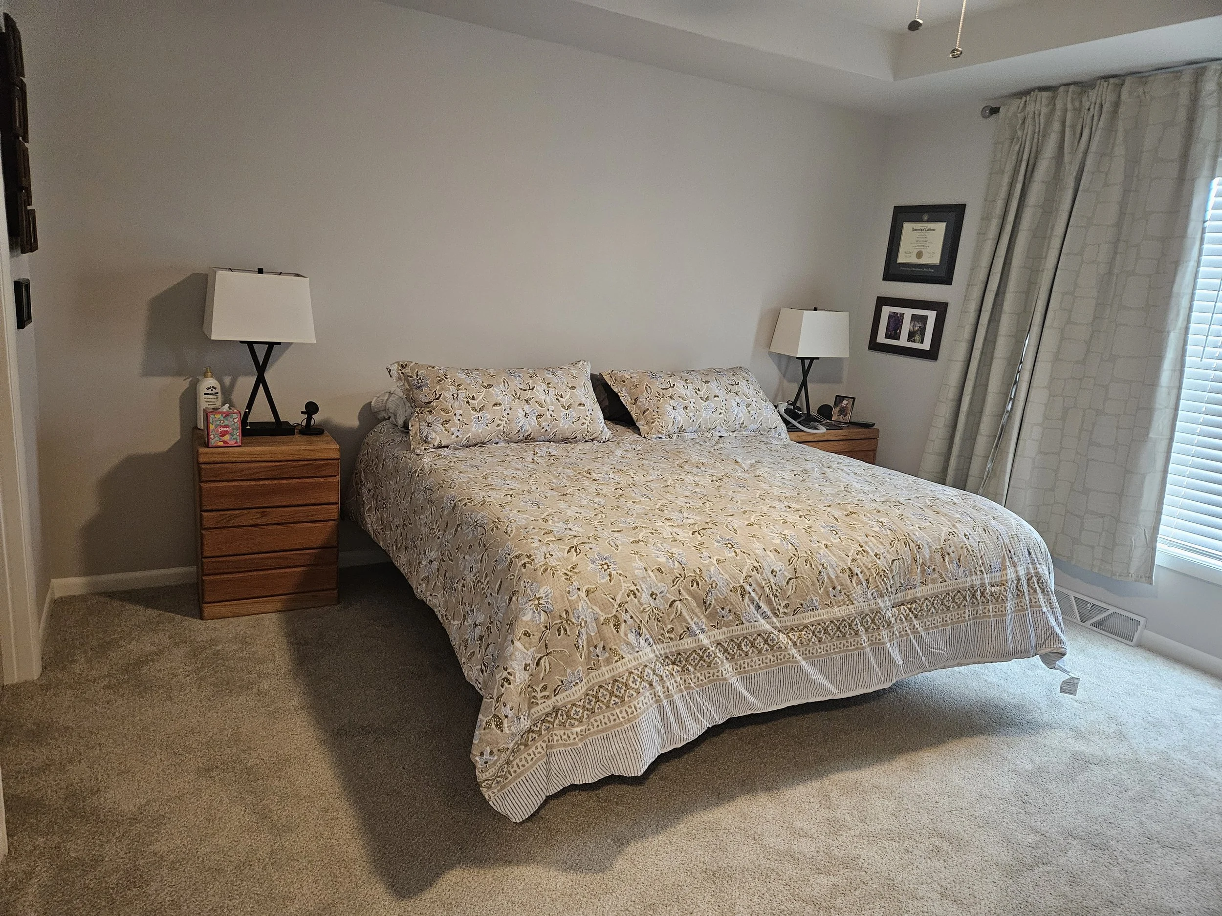 A bedroom with a bed covered in a floral bedspread, two matching pillows, and two nightstands with lamps on either side. One nightstand has a hand sanitizer and a small colorful item, and the other has picture frames and a phone. There are framed pictures on the wall and closed blinds covering a window with curtains, and beige carpet on the floor.