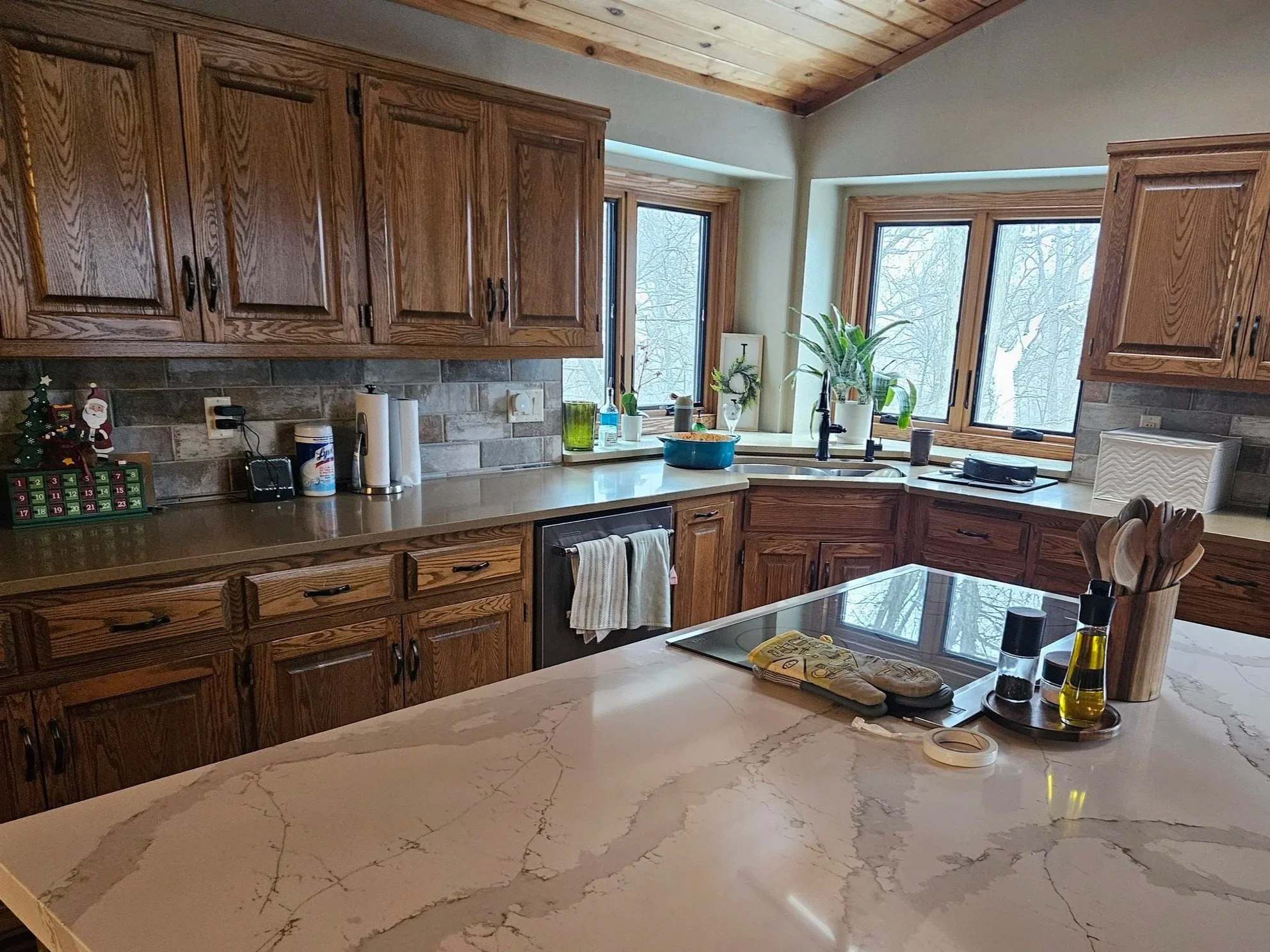 A kitchen with wooden cabinets, marble countertops, and large windows overlooking a snowy outdoor scene. There are various kitchen items on the counter including potted plants, a dish drying rack, and a tray with spices, while the island has utensils, a salt and pepper shaker, and olive oil.