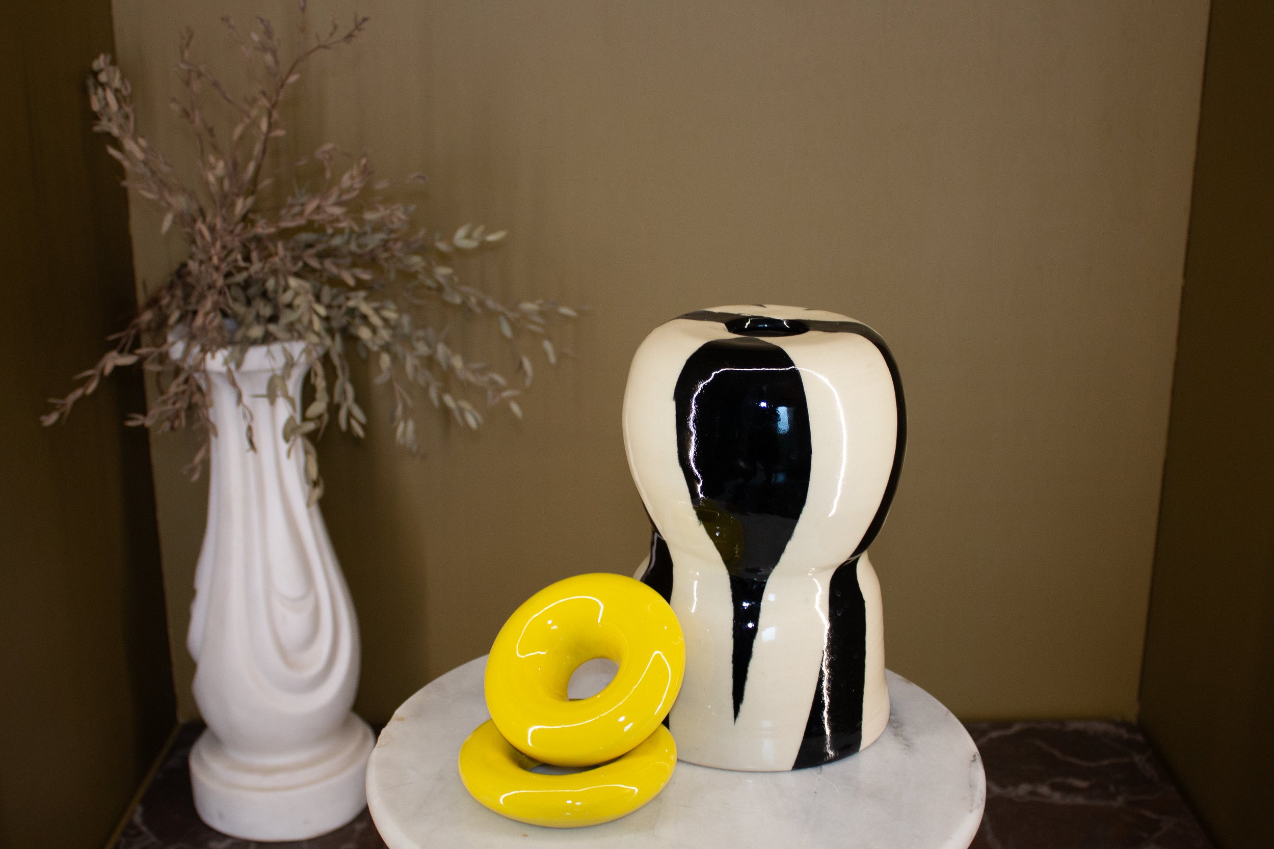 A decorative arrangement featuring a black and white stripped ceramic sculpture, and a yellow doughnut shaped ceramic piece on a white marble table against a green wall.