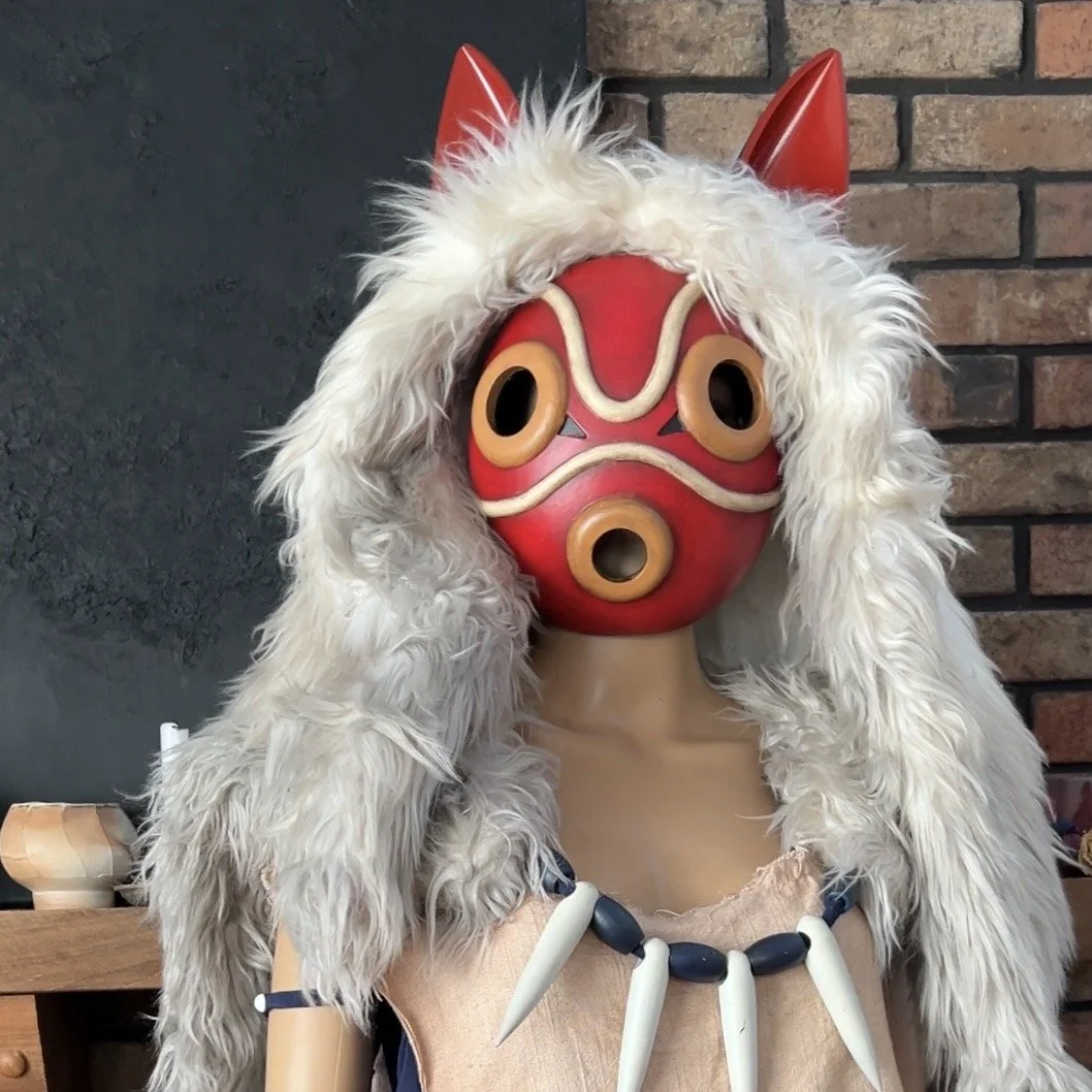 Person wearing a red and beige mask with a fur hood, resembling a character from Japanese folklore, and a necklace with large white teeth or bones, standing in front of a brick wall.