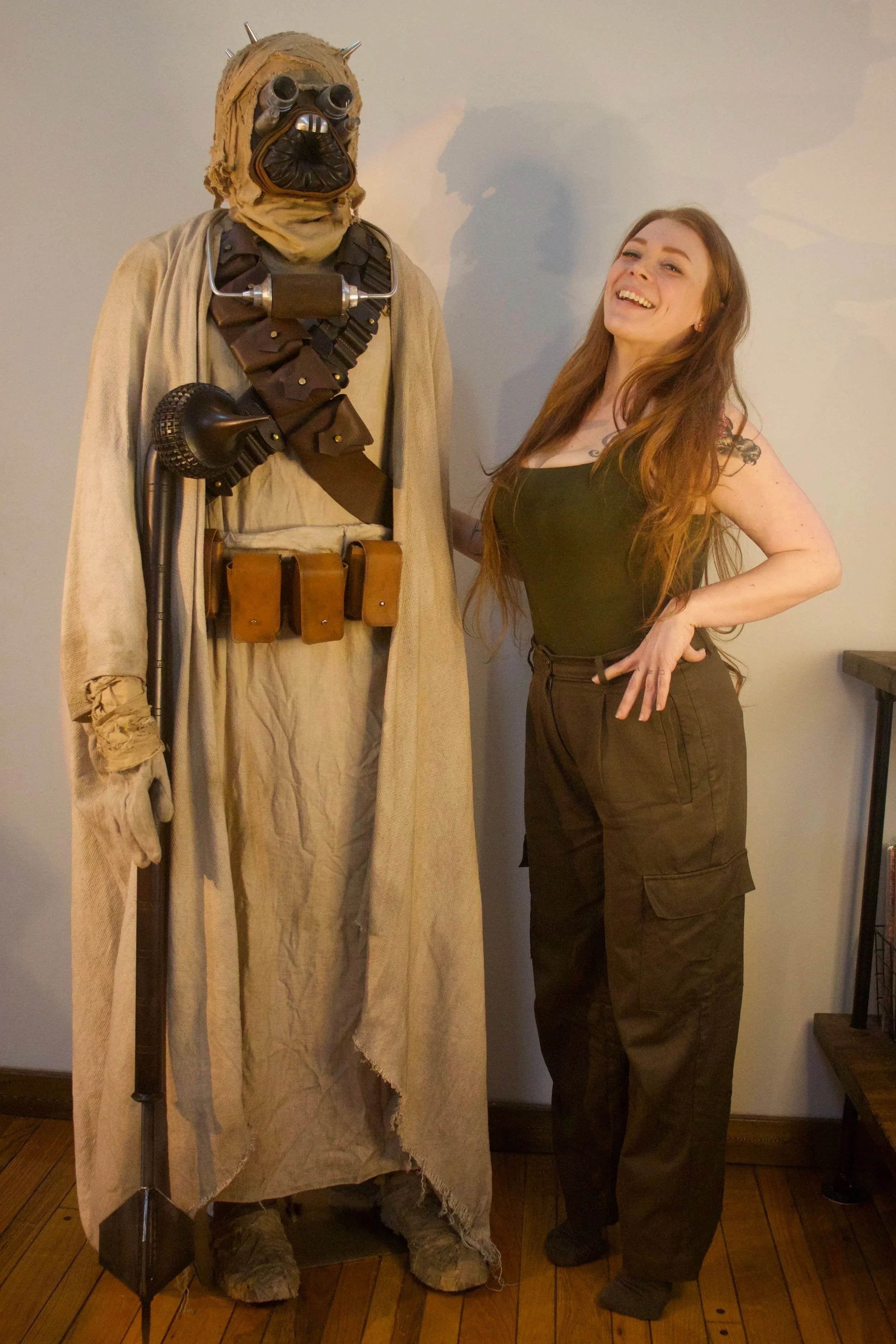 A woman with long red hair standing next to a life-sized Threepio and Chewie Star Wars character made of costumes and props, inside a room with wooden floors and a light-colored wall.