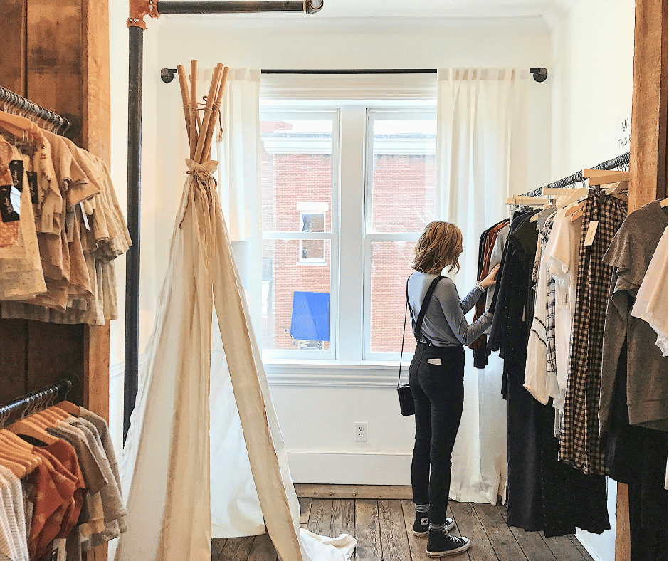A woman shopping in a boutique clothing store, looking at a rack of clothes near a window.