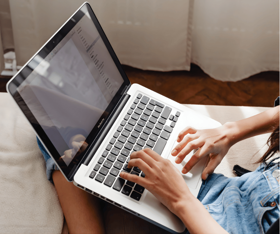 Person using a MacBook Air laptop on their lap, sitting on a couch with a curtain in the background.
