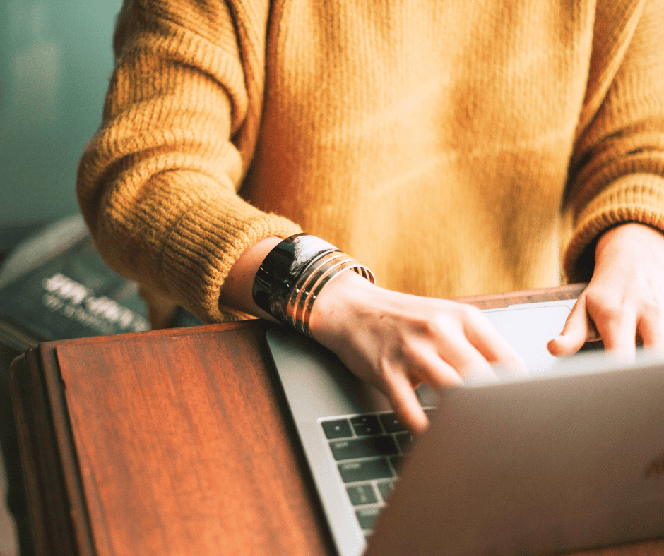 Person wearing a mustard-colored sweater and shiny black and metallic bracelets typing on a silver laptop at a wooden desk, with a closed book in the background.