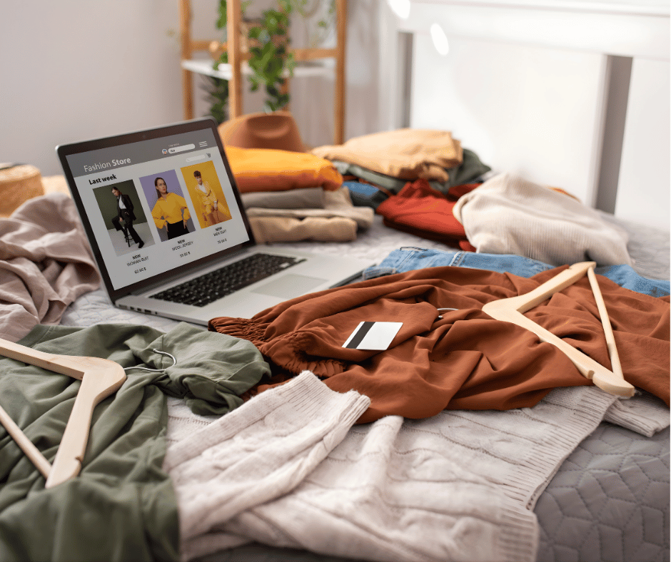 A cluttered bed with various pieces of clothing, a laptop displaying a fashion store website, and some empty wooden hangers.