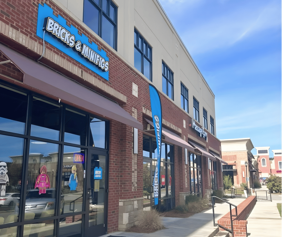 Exterior view of a brick storefront with a sign that reads 'Bricks & Minifigs' and large windows displaying LEGO characters inside. There are awnings, a blue display flag, and a sidewalk with a railing in front of the store. The sky is blue with some clouds.