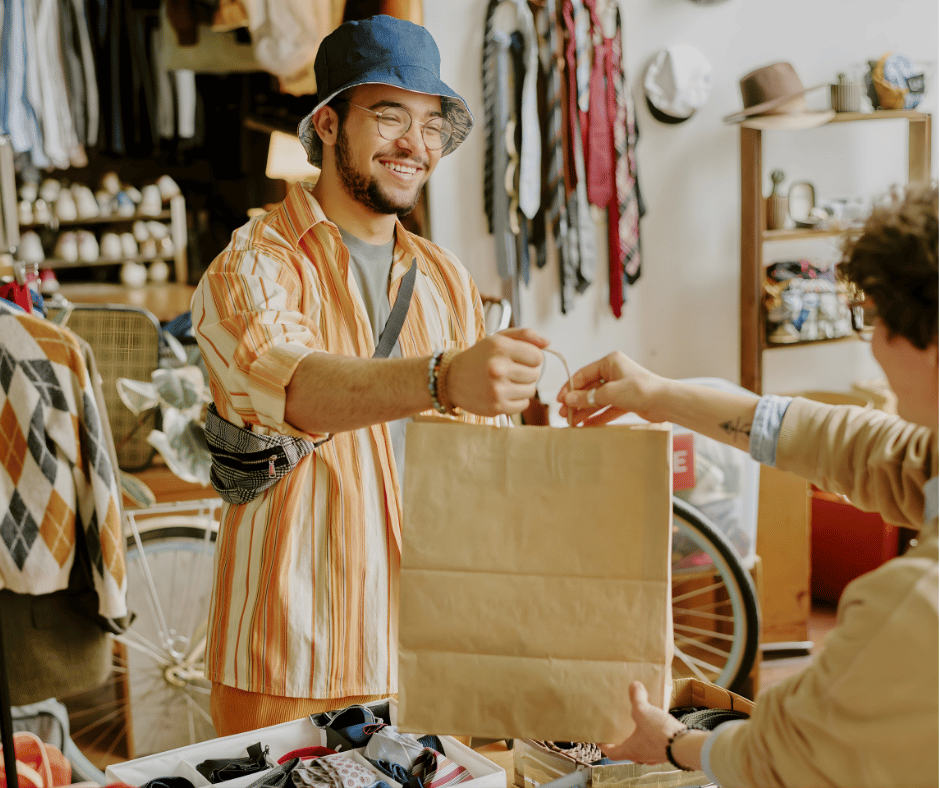 A smiling man wearing glasses, a blue bucket hat, and a striped shirt is exchanging a paper shopping bag with another person inside a thrift store or flea market. They are surrounded by clothing and accessories on display.