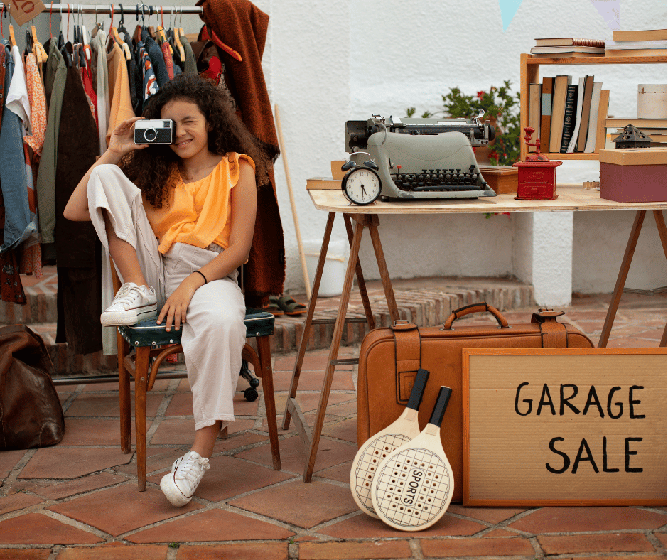 A girl with curly hair sitting on a chair at a garage sale, taking a photo with a camera. There are clothing racks, vintage items, books, and sports rackets around her.