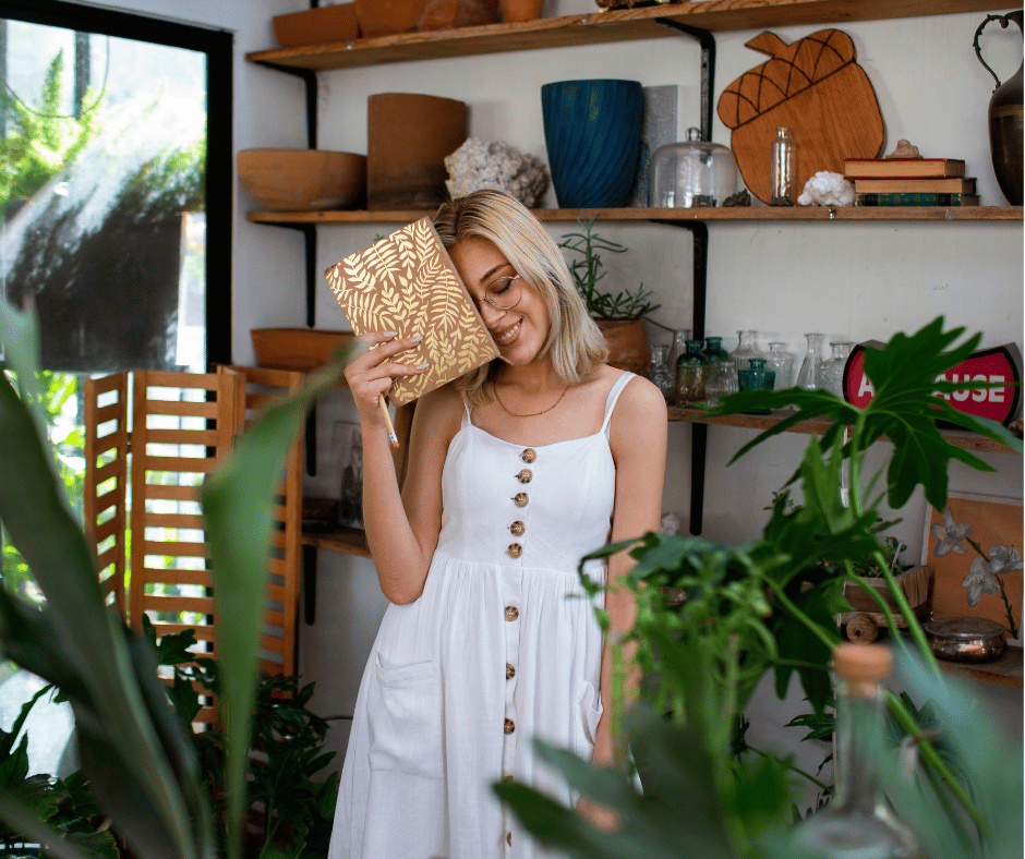 A woman with blonde hair wearing a white dress and glasses, holding a patterned book to her head and smiling, standing in a room with wooden shelves and plants.