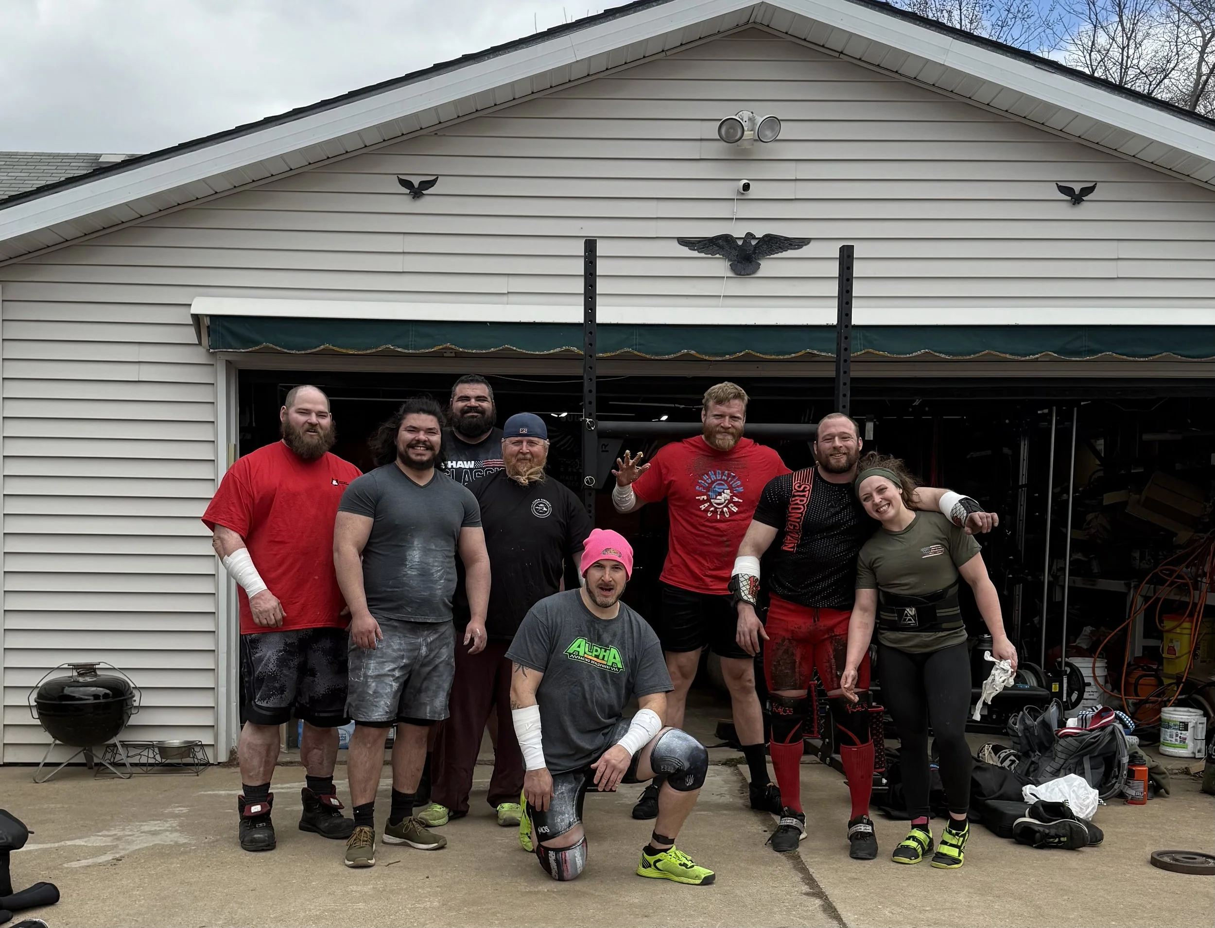 Group of seven people, six men and one woman, standing outside a garage, some with injury wraps, smiling after a workout or competition.