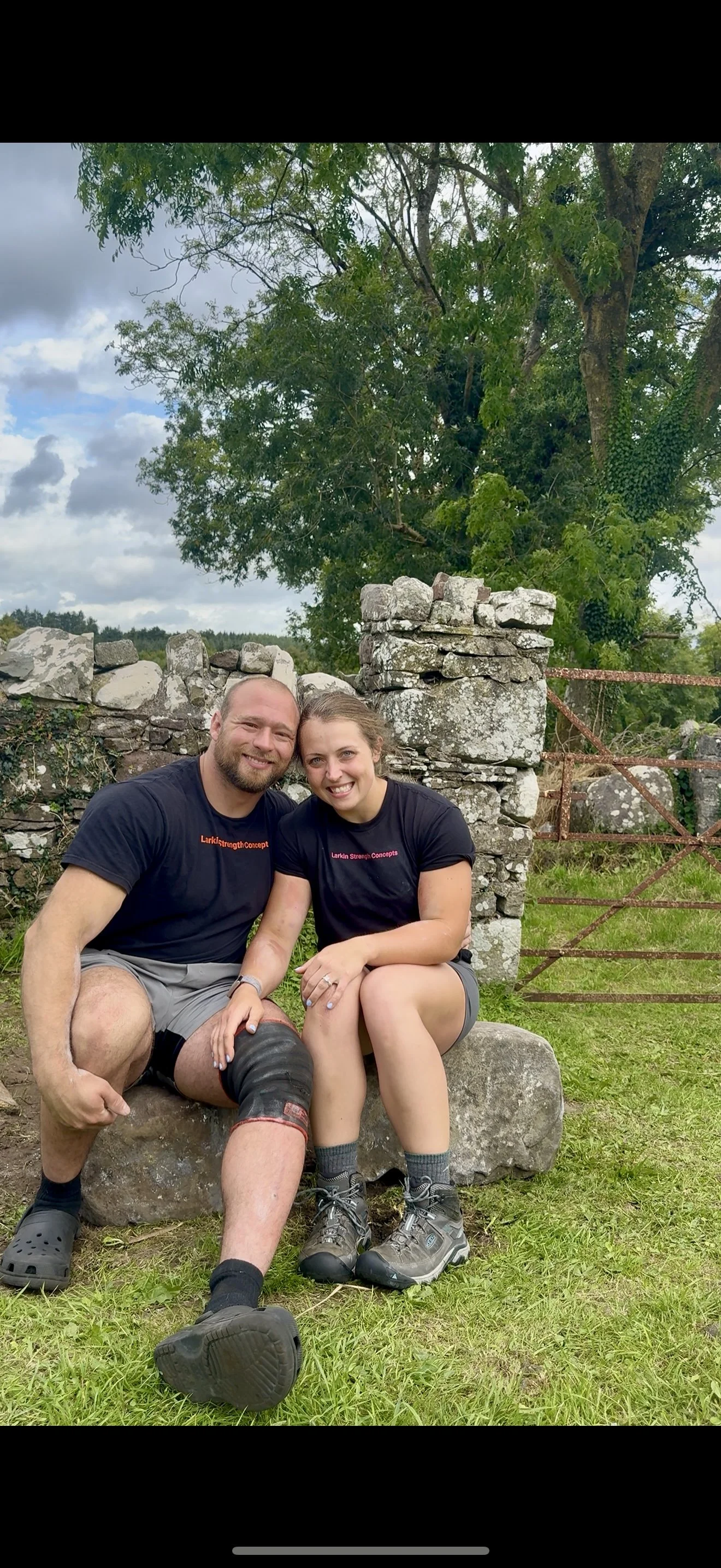 A smiling man and woman sitting on a stone bench outdoors in front of a stone wall and green trees. They are dressed in casual outdoor clothing, with the man wearing a black T-shirt and shorts, and the woman wearing a black T-shirt and shorts. The man has a knee brace on his left leg.