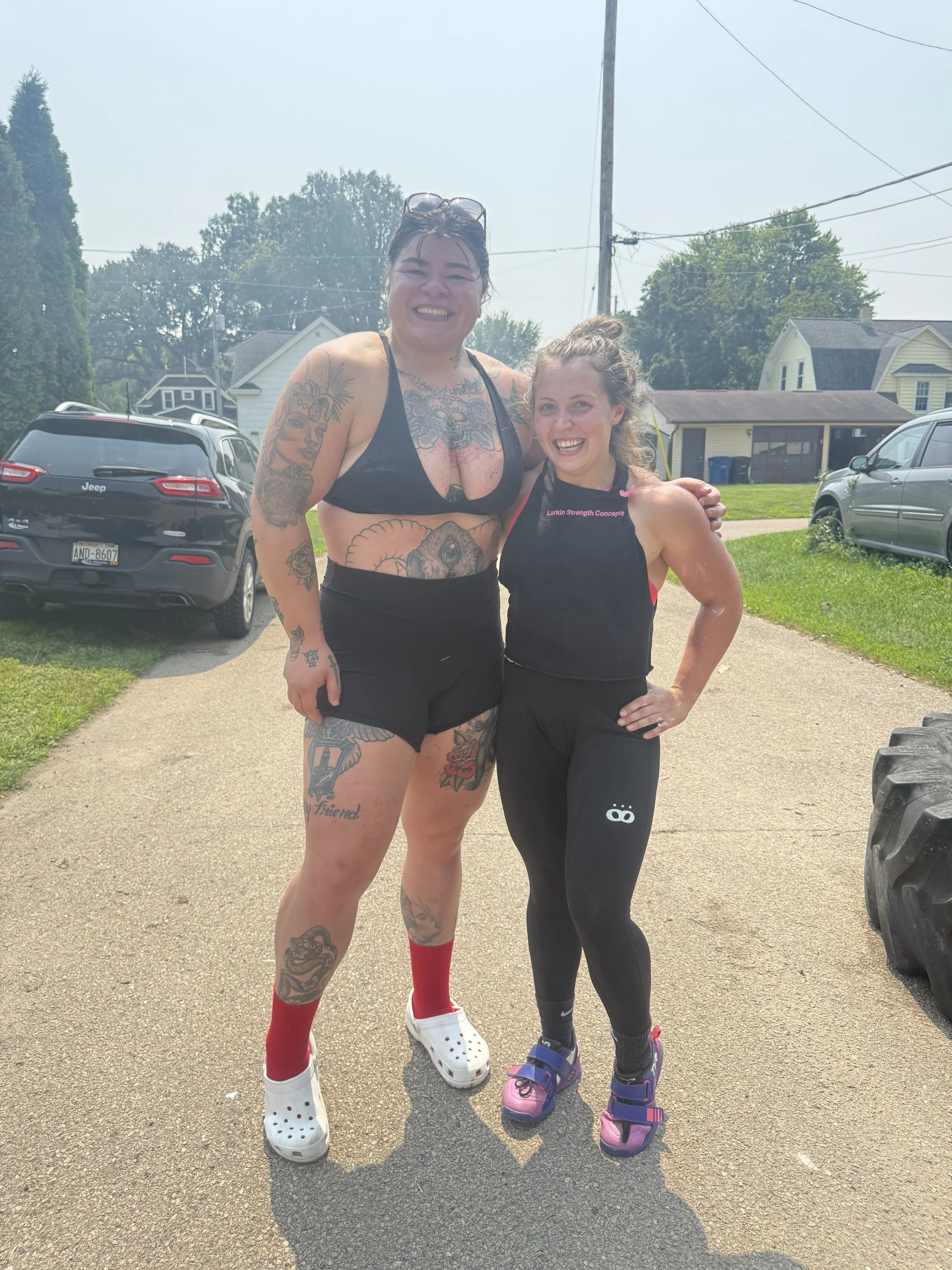 Two women, one with tattoos and in casual workout attire, and another in workout clothes, standing outside on a driveway, smiling and posing after exercising.