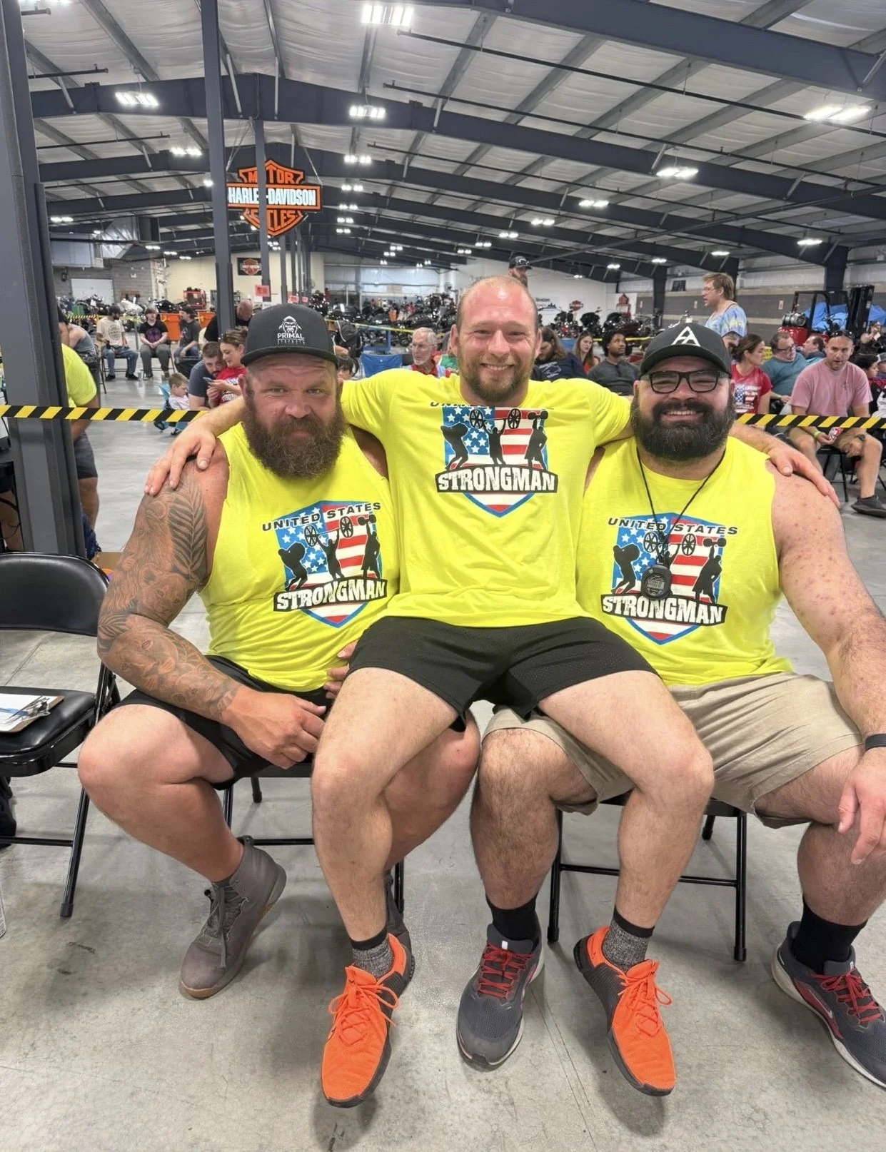 Three men wearing bright yellow shirts with 'United States Strongman' logo, sitting closely together and smiling at a strongman event inside a large warehouse, with motorcycles and people in the background.