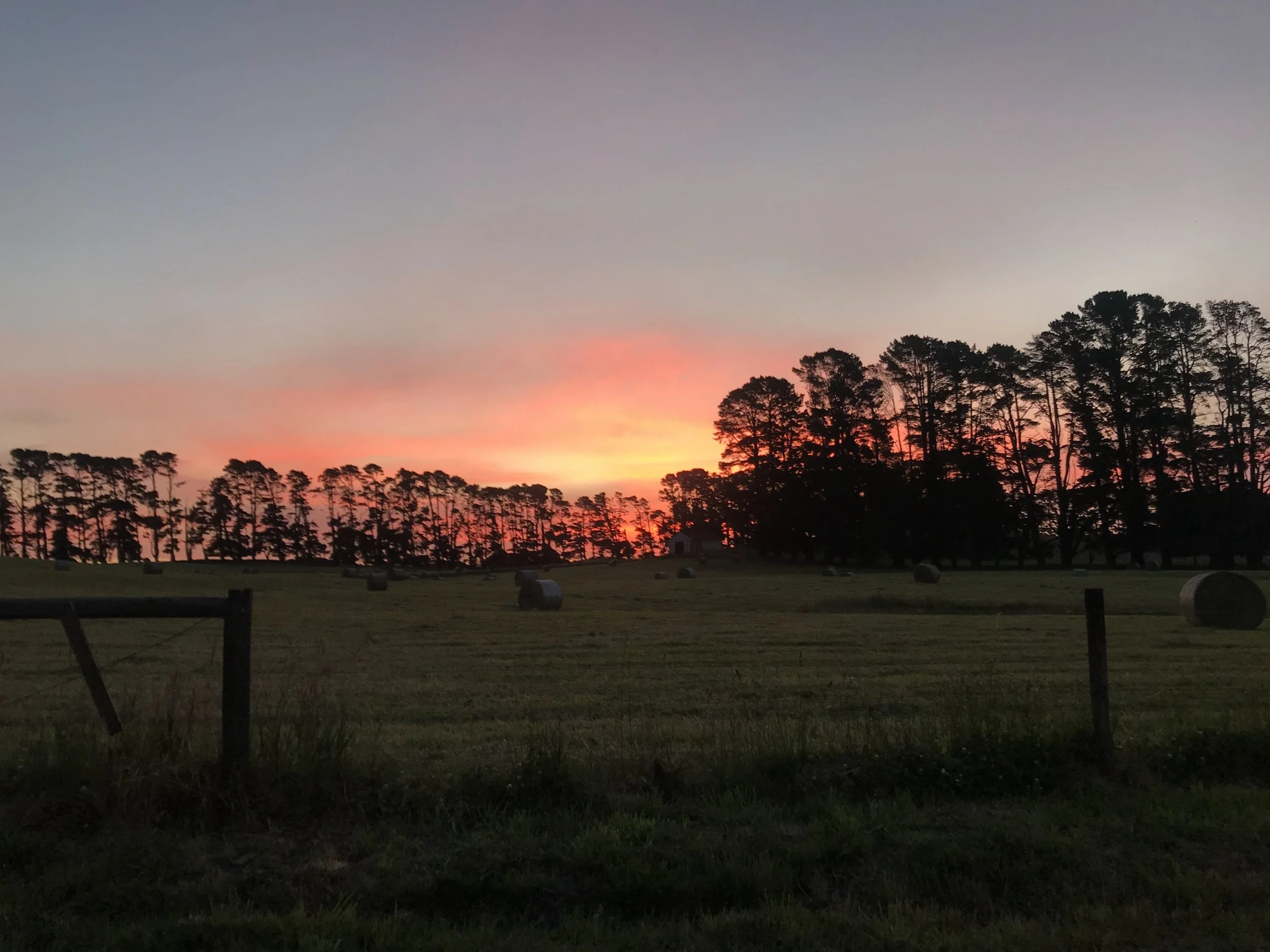 A rural landscape at sunset with a colorful sky, silhouette trees on the horizon, and hay bales scattered across a grassy field, with a wooden fence in the foreground.