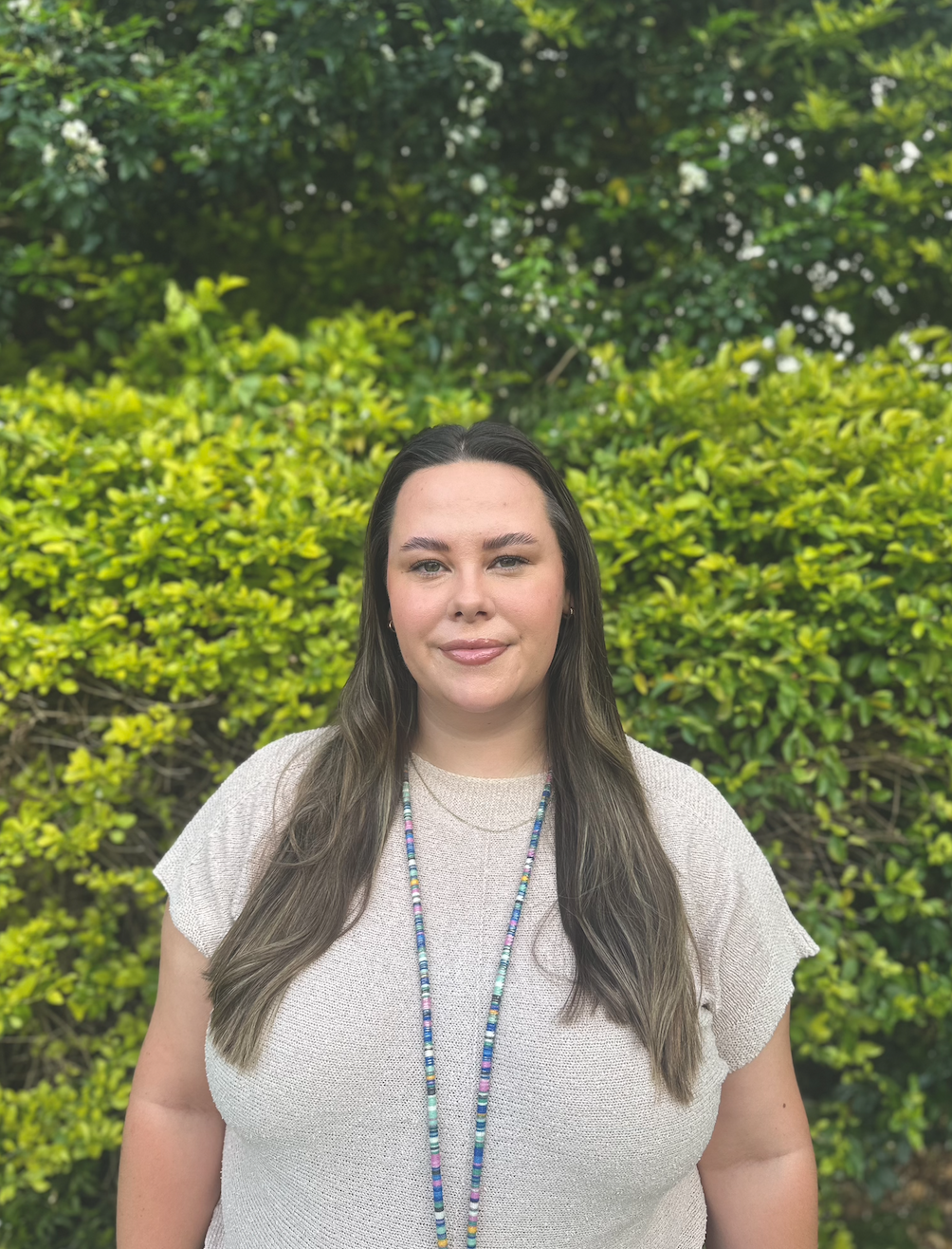 A woman with long dark hair and light skin standing outdoors in front of lush green bushes and trees, wearing a beige shirt and a colorful beaded necklace.