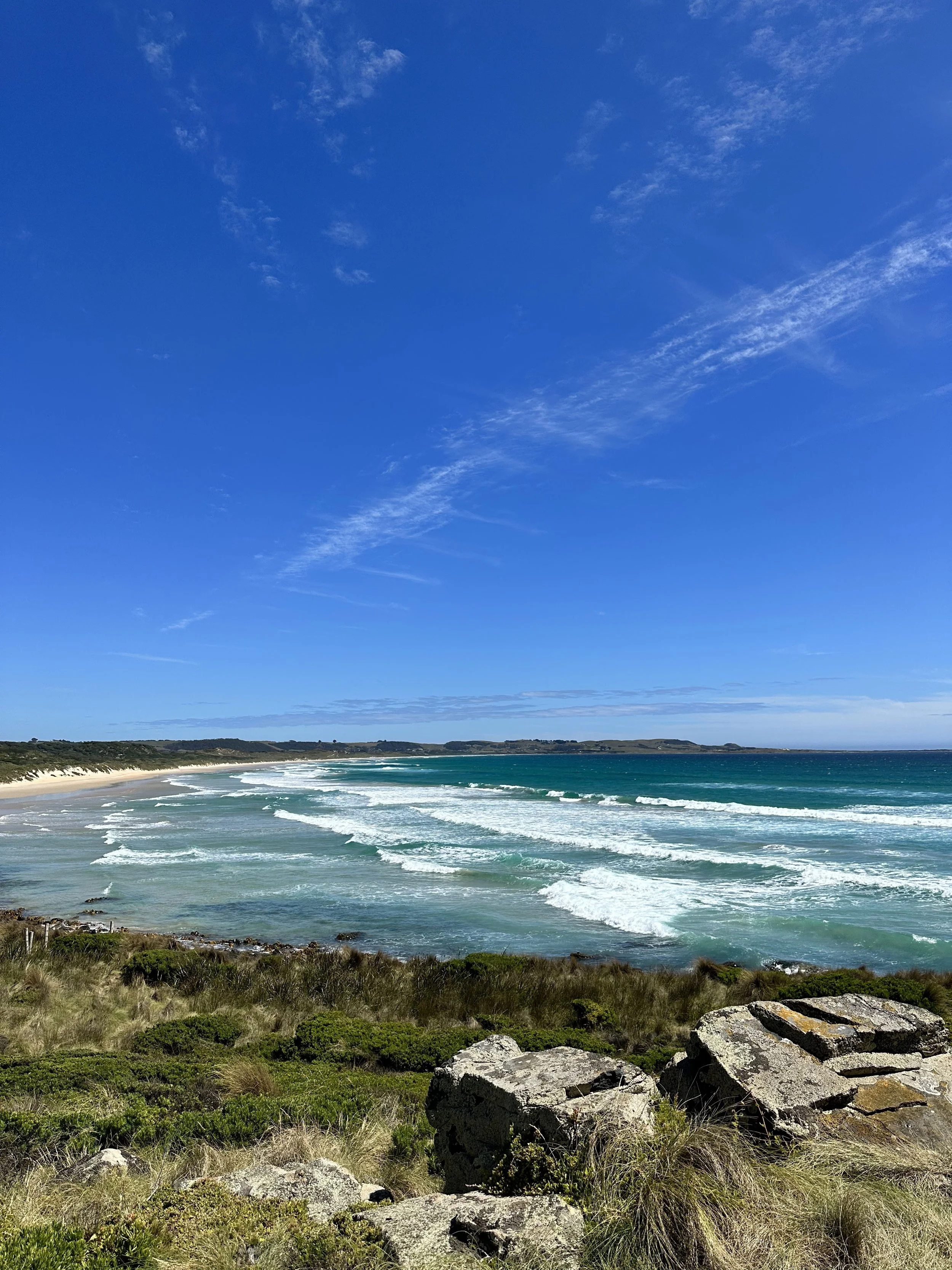 A scenic view of a beach with waves crashing onto the shore, under a bright blue sky with some clouds.
