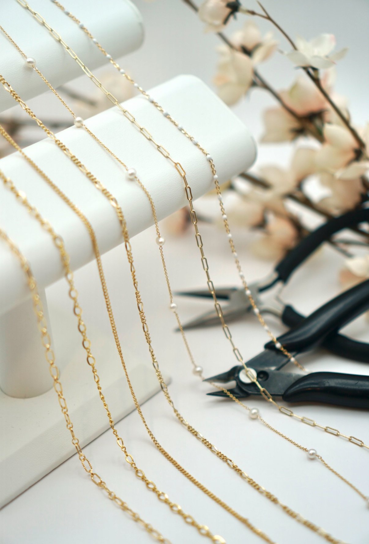 Gold and sterling silver permanent jewelry chains with pearls displayed on a jewelry stand, with a pair of black pliers and artificial flowers in the background.