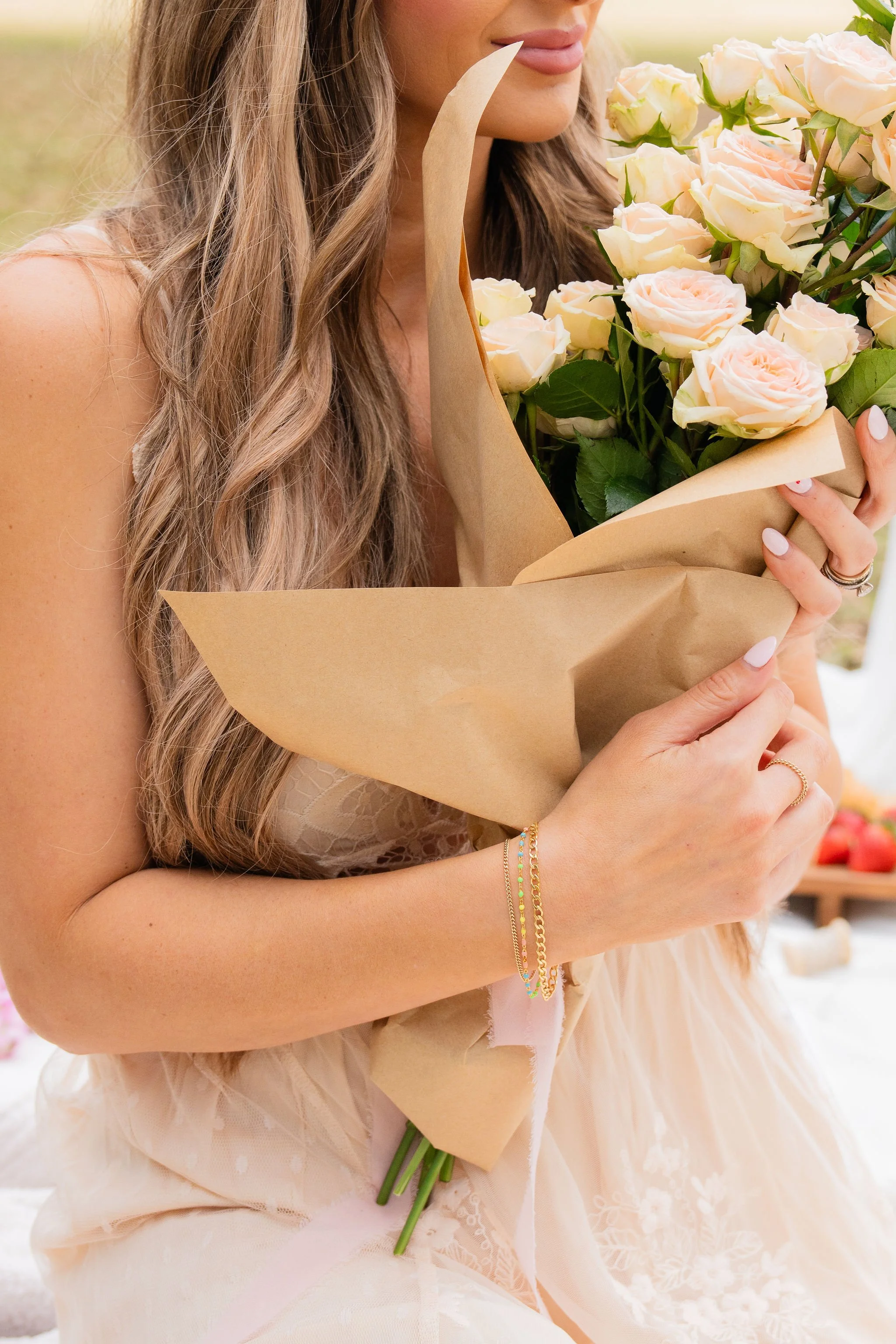 A woman with long, wavy hair holding a bouquet of white and light pink roses wrapped in brown paper, wearing a light-colored dress and jewelry.
