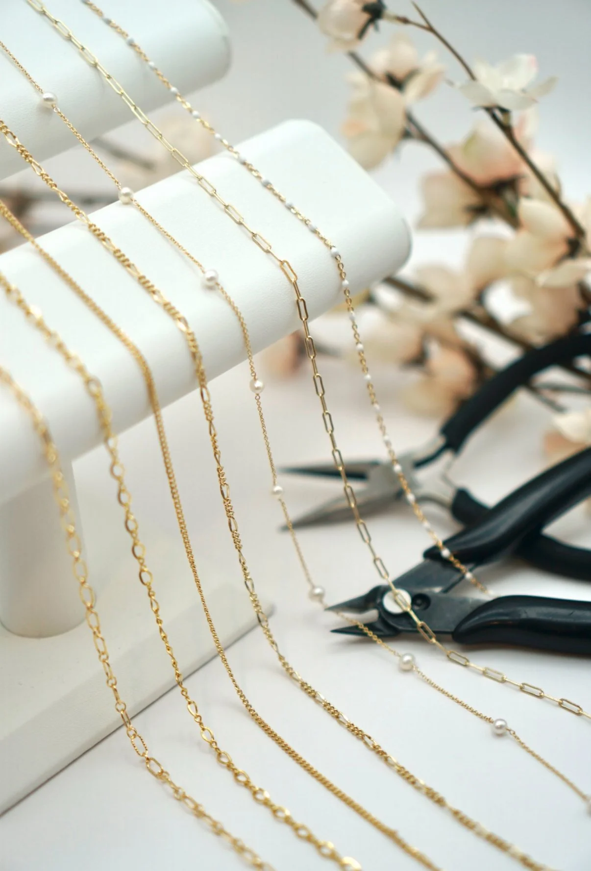 Gold and silver necklaces with various chain styles, some adorned with small pearls, displayed on a white jewelry stand with a pair of black jewelry pliers nearby and soft pink flowers in the background. Handcrafted. Welded. Permanent.