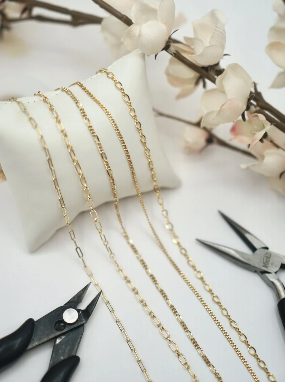Permanent jewelry gold chains displayed on a white jewelry pillow with pliers nearby, alongside a branch of light-colored flowers in the background.