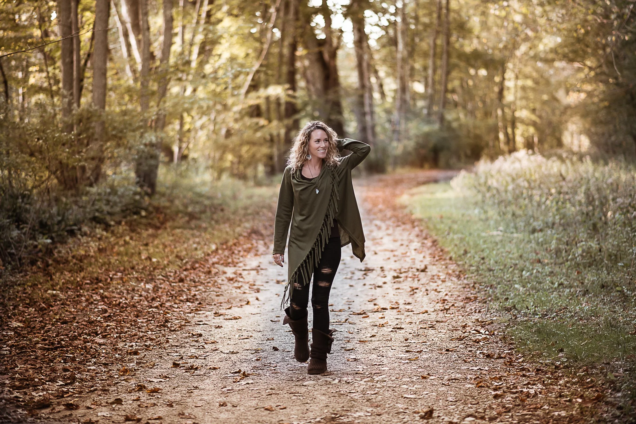 About the Jeweler herself; a woman with curly hair walking along a leaf-covered trail in a forest during autumn, smiling and looking to her left, wearing a green jacket, black ripped jeans, and brown boots. Beach, Jewelry, Artistan, Earrings