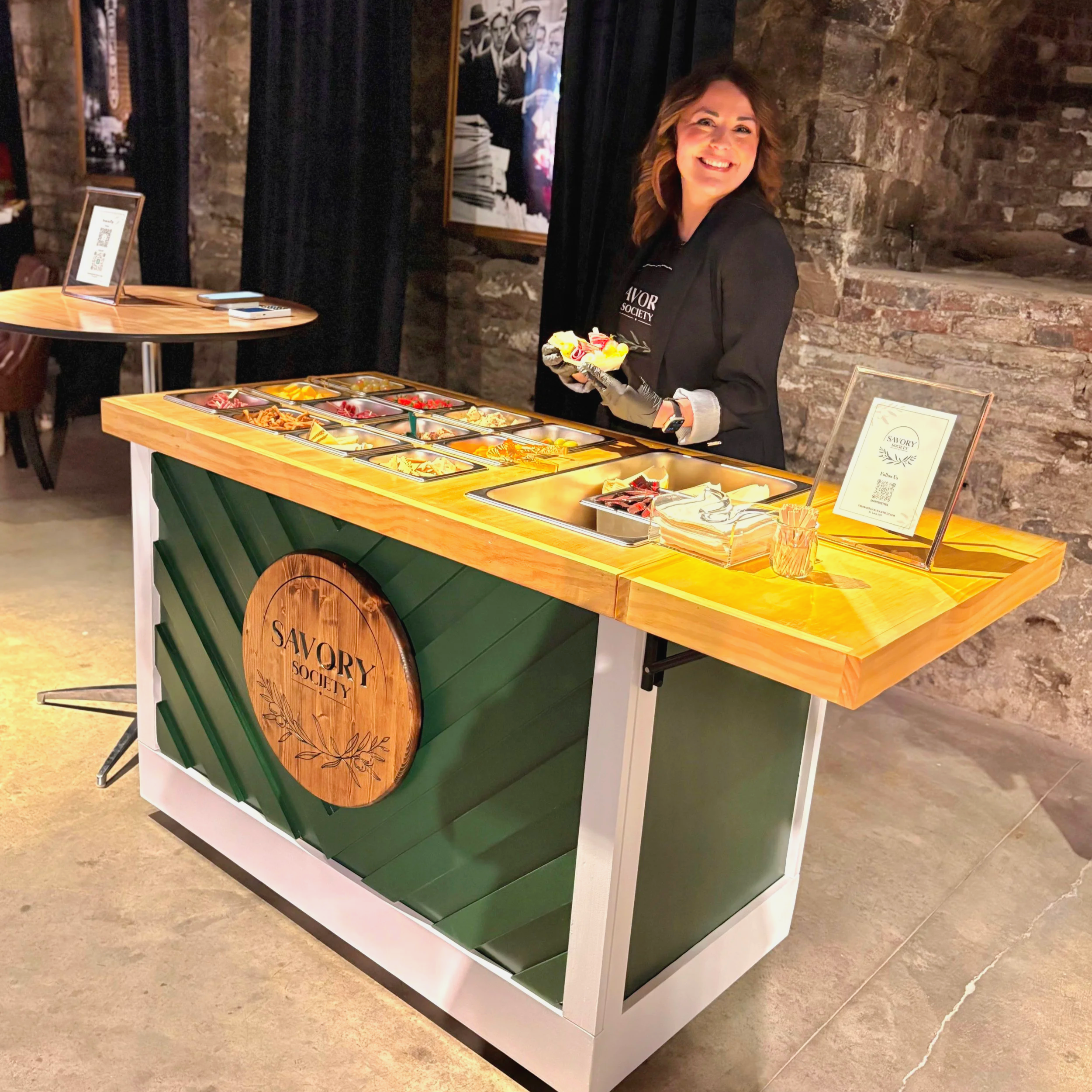 A woman Standing behind a mobile charcuterie cart with various food items, wearing a black jacket and gloves, in a wedding venue setting with brick walls.