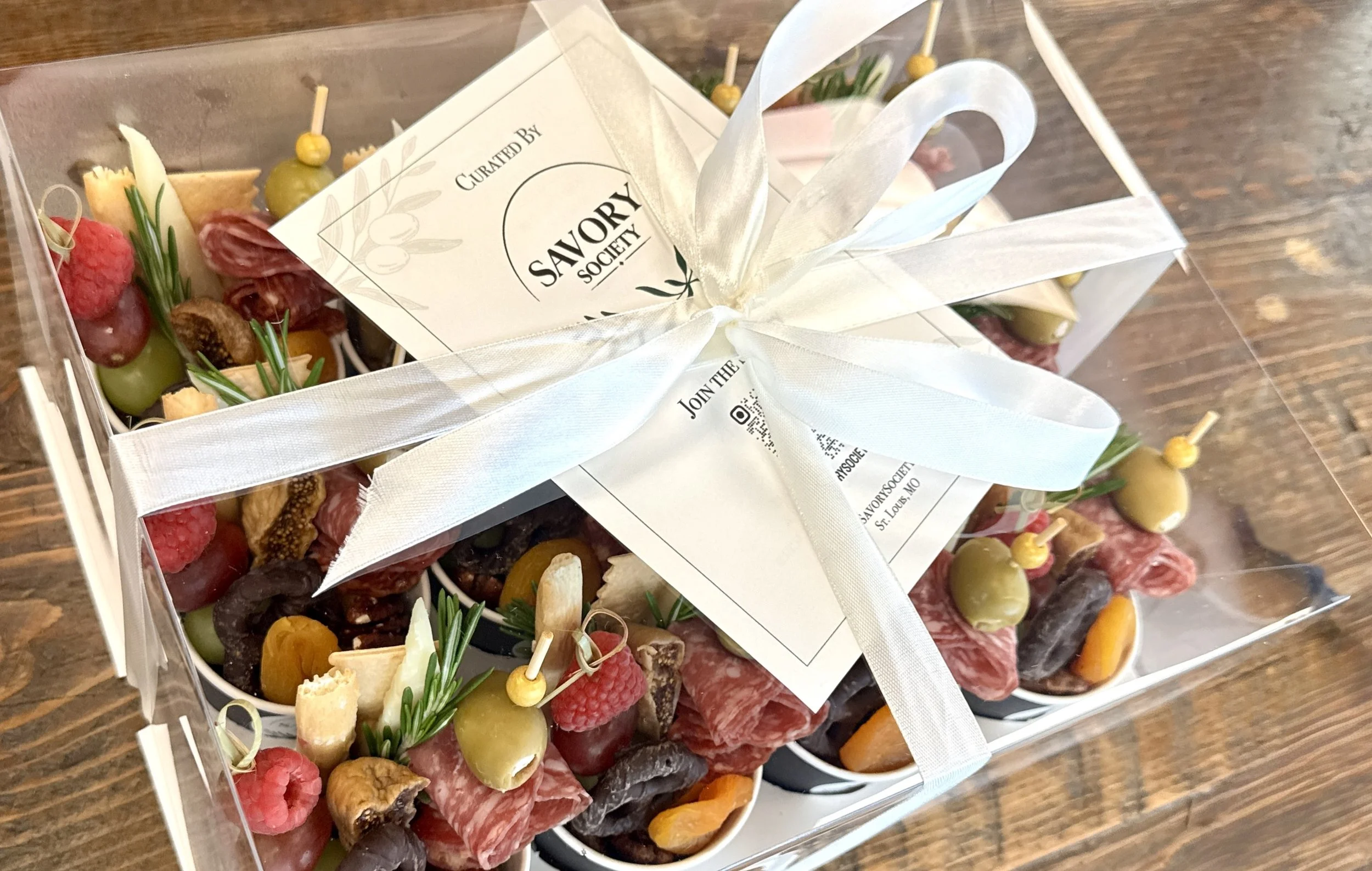 A gift box containing assorted cheeses, meats, crackers, grapes, olives, and garnishes, wrapped with a white ribbon and a note from Savory Society.