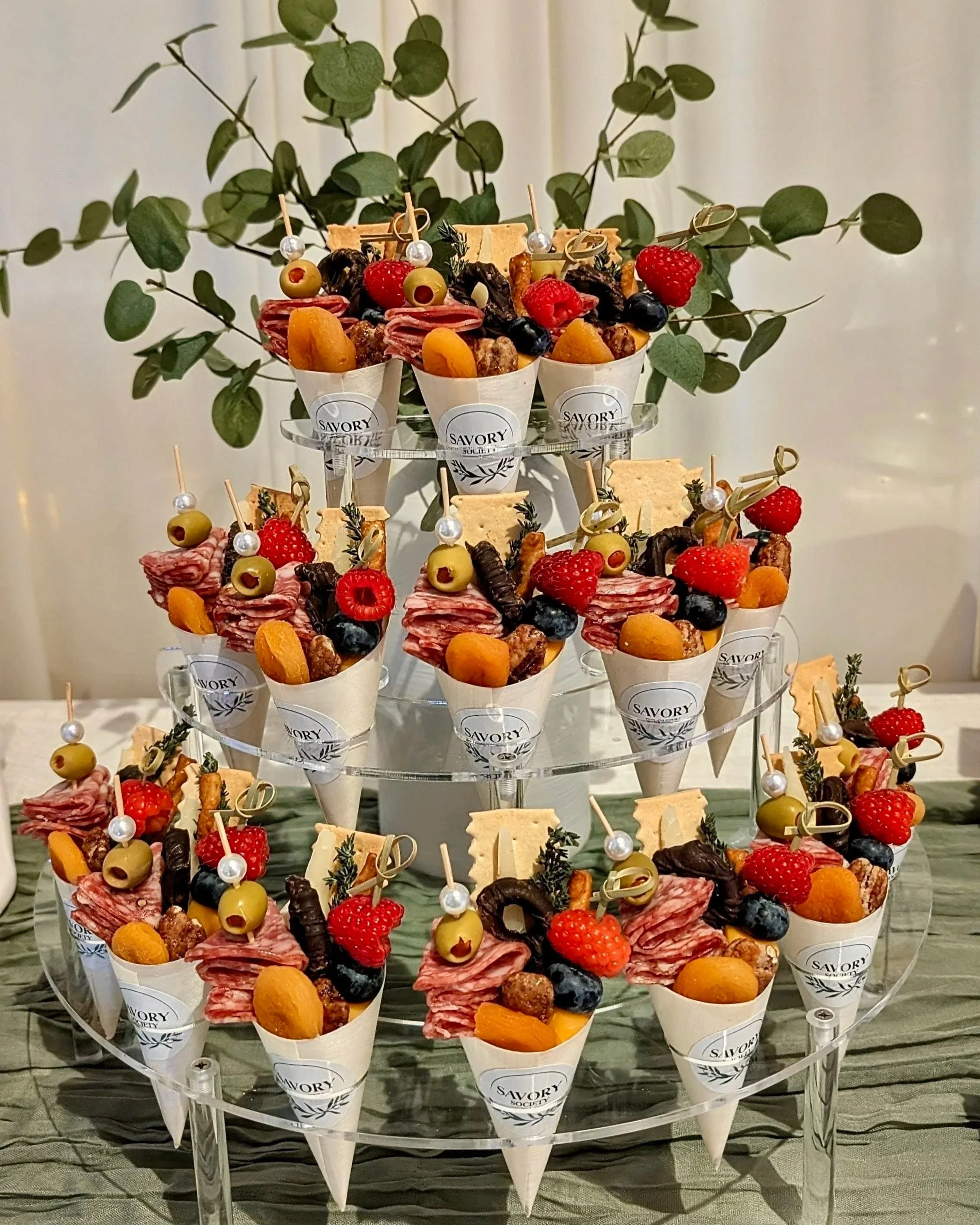 Tiered display of assorted cheese, meat, and fruit skewers in cone-shaped cups labeled 'Savory Society' with a green leafy plant in the background.