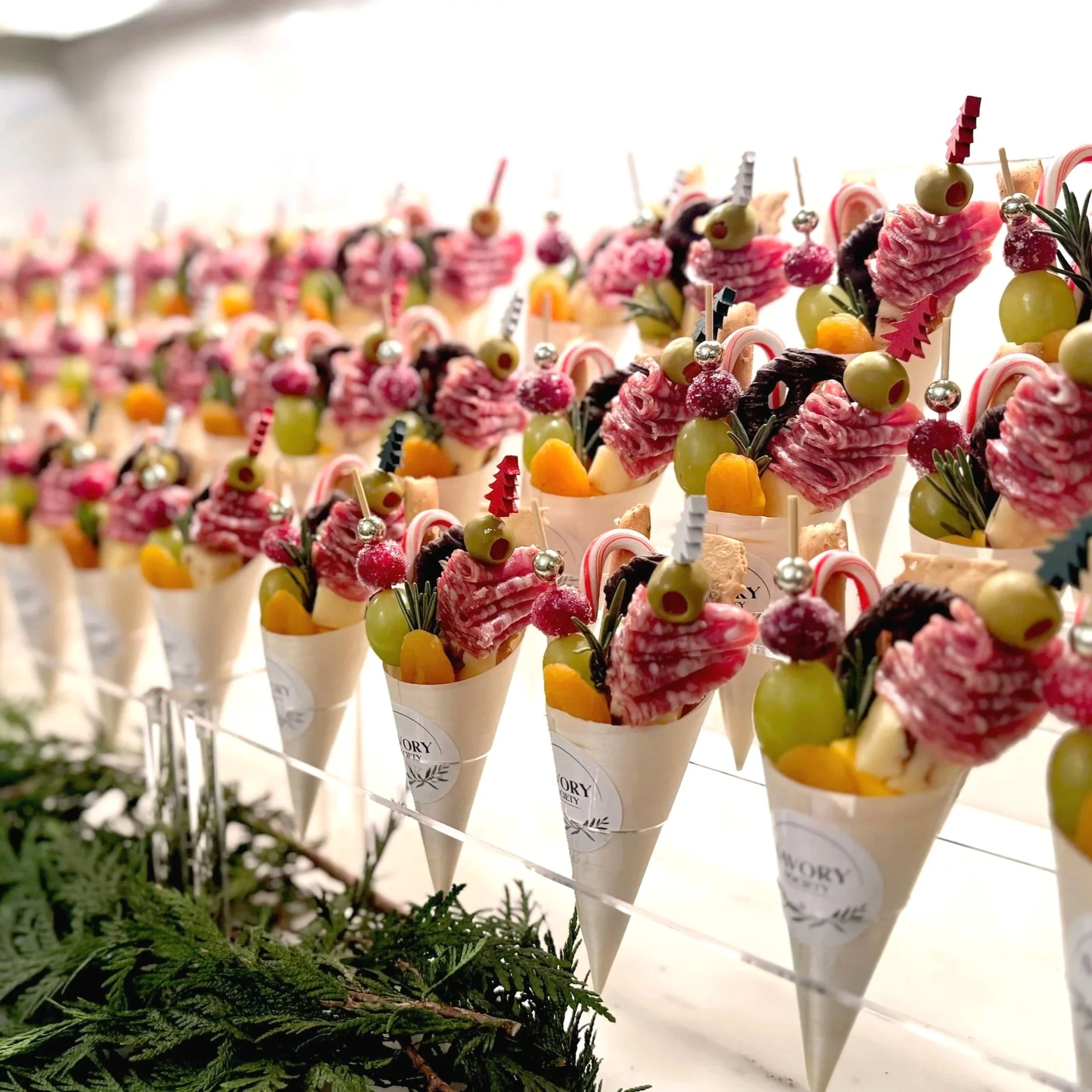 Assorted charcuterie cones decorated with fruits, candies, and herbs, displayed on a white surface.