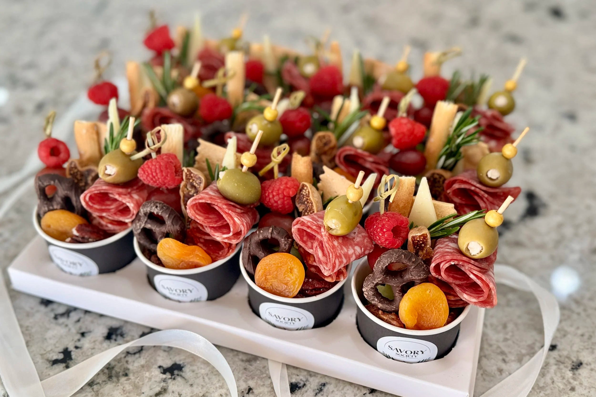 Gift box of assorted charcuterie cups with cured meats, cheese, olives, raspberries, apricots, and rosemary sprigs on a white platter.