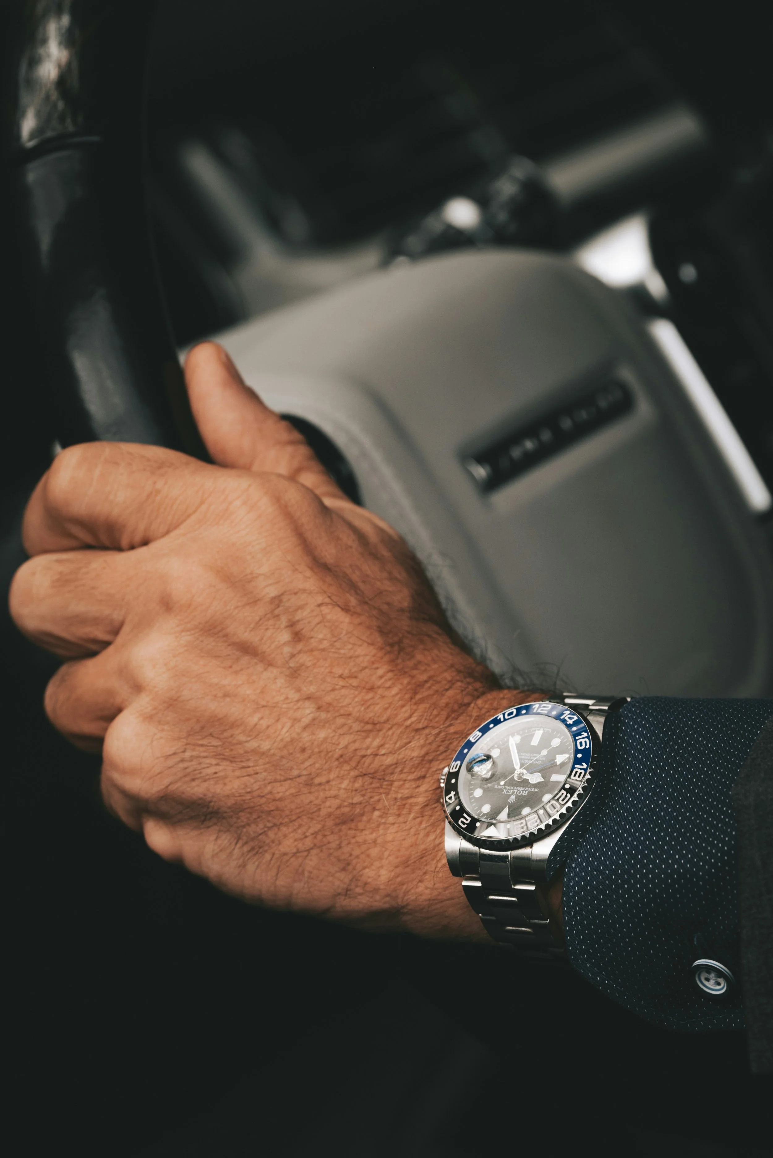 Close-up of a person's left hand on a steering wheel, wearing a silver and black wristwatch with a blue bezel, inside a vehicle.
