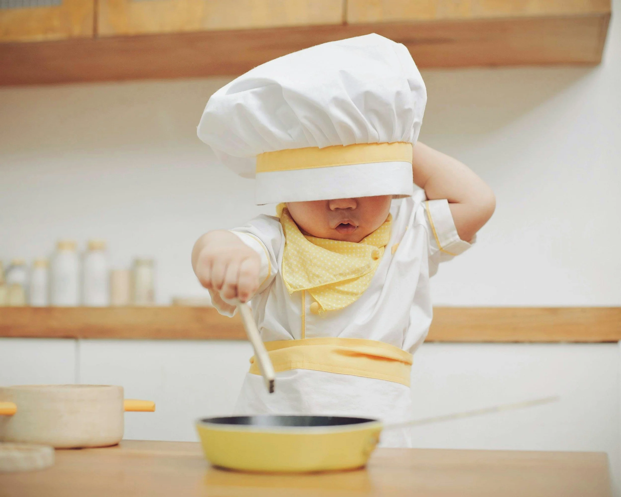 Young child dressed as a chef, wearing a large white chef hat, yellow apron, and a yellow bib, stirring a small pan in a kitchen setting.