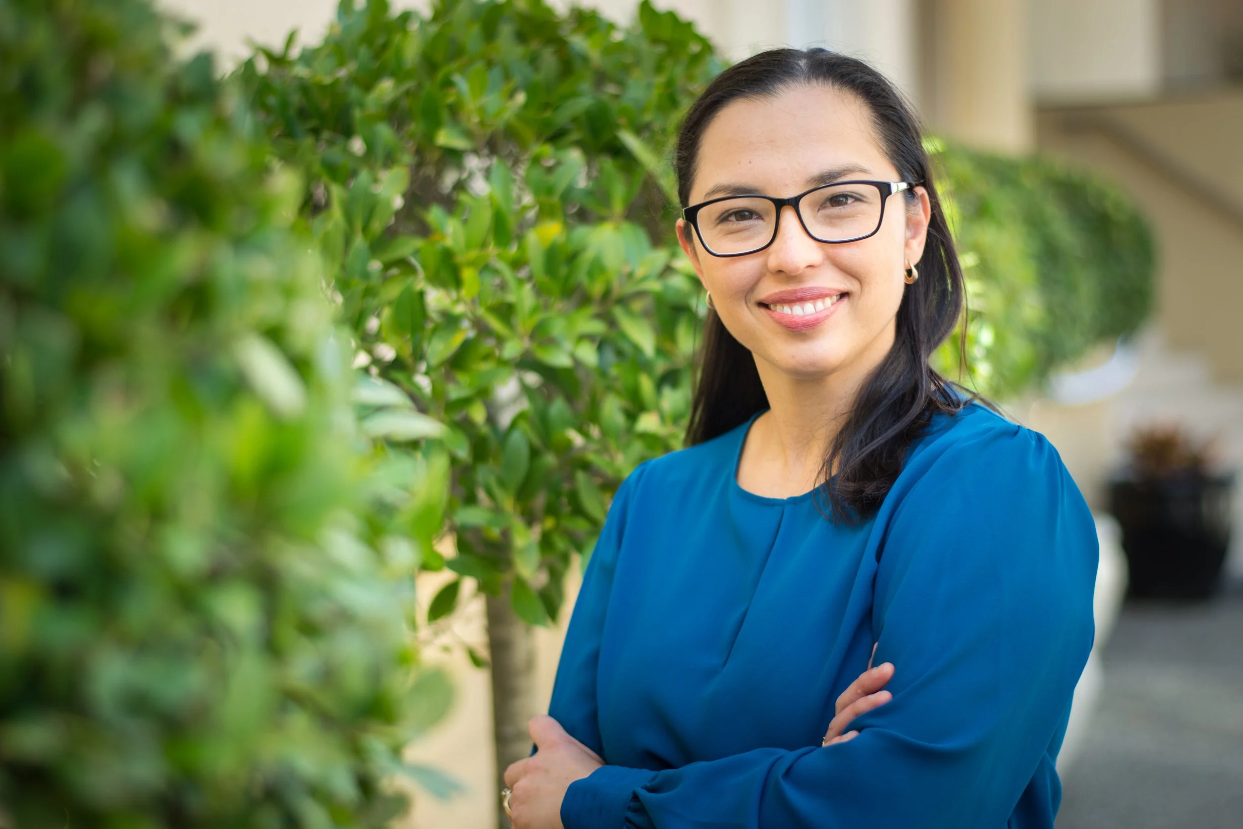 A woman with black hair and glasses smiling outdoors with green shrubbery in the background.