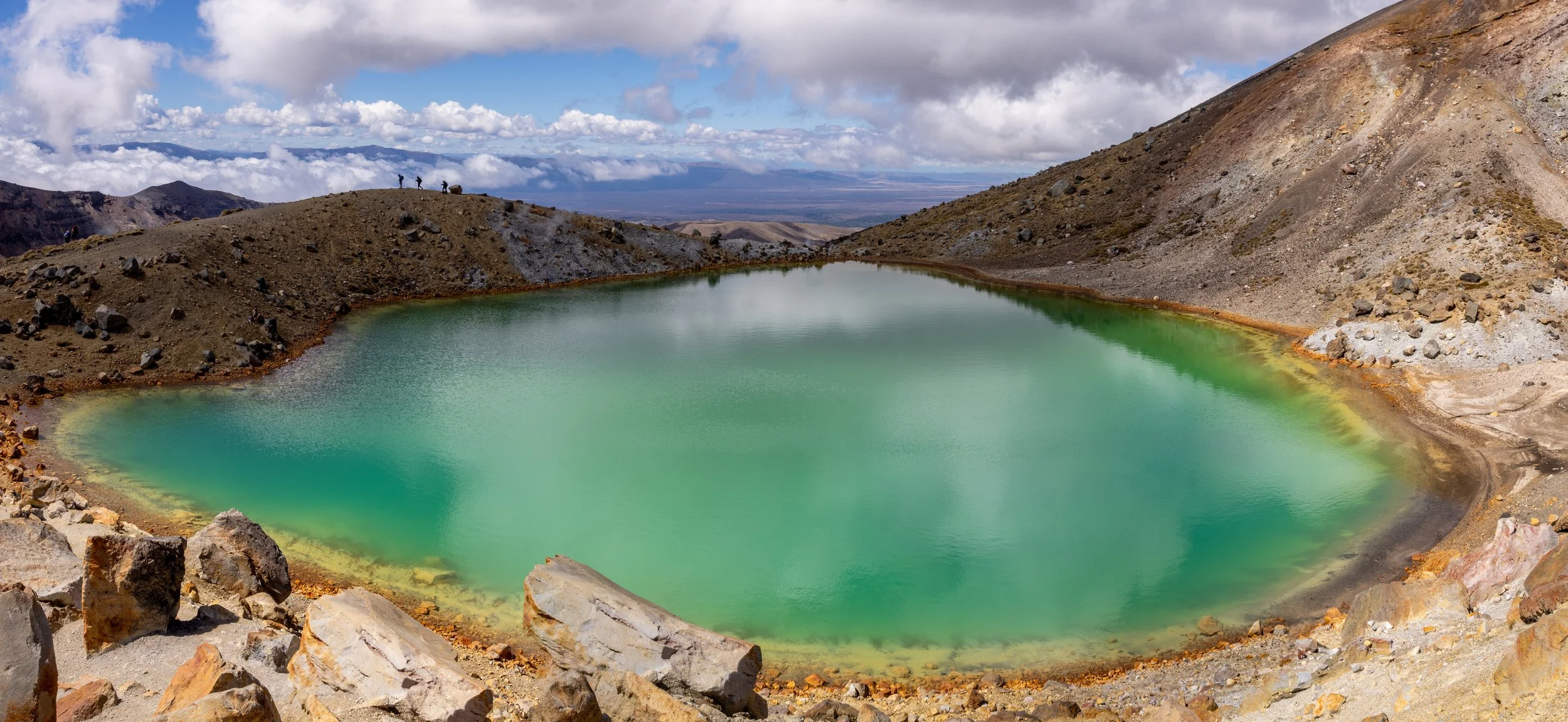 A crater lake with green water, surrounded by rocky, barren volcanic landscape under a partly cloudy sky.