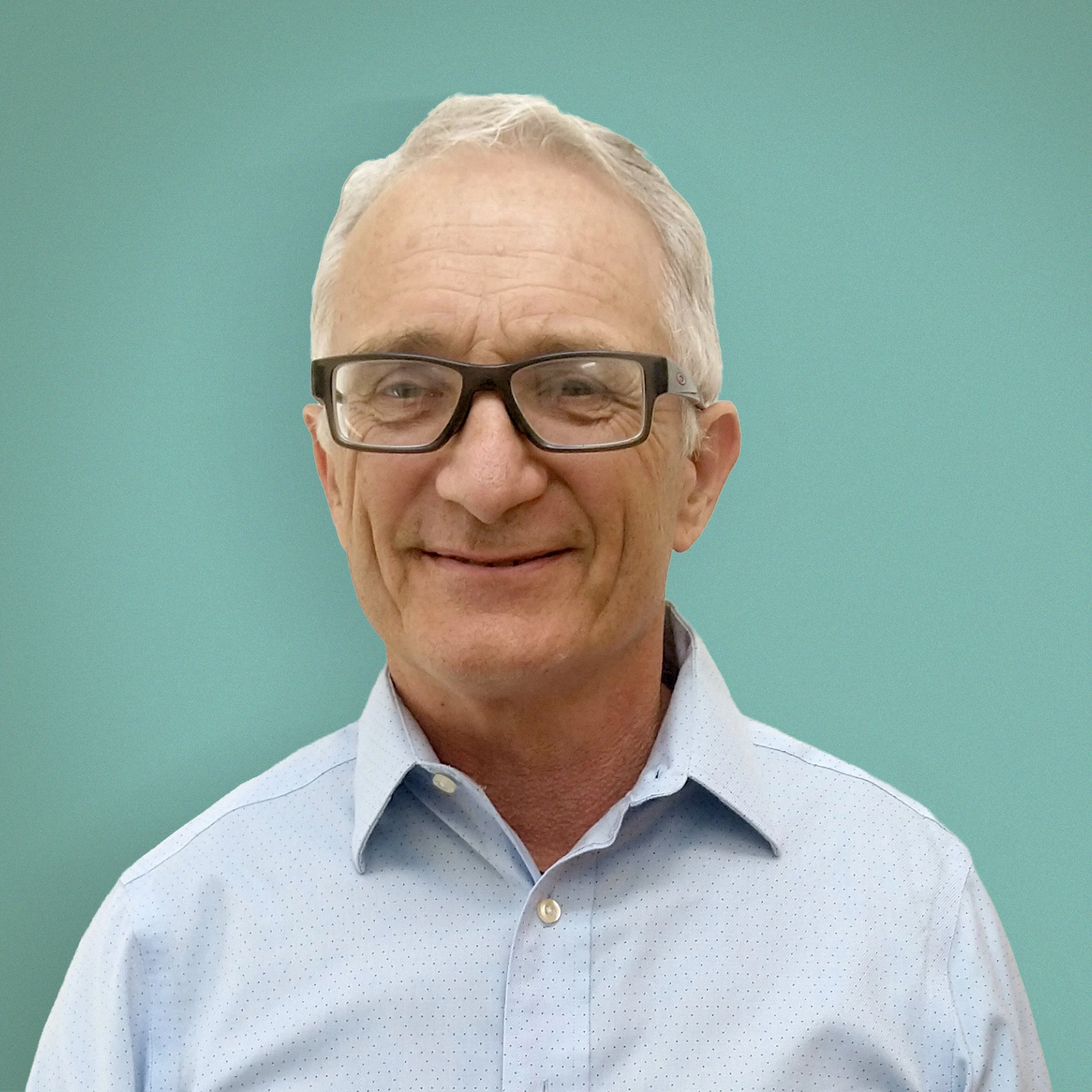 A middle-aged man with gray hair, glasses, and a light blue collared shirt smiling against a teal background.