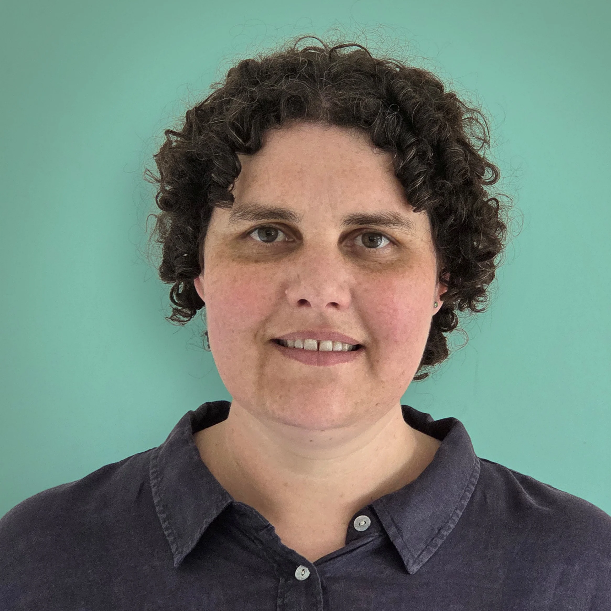 A woman with short curly dark hair, wearing a dark collared shirt, standing in front of a plain, light-green background.