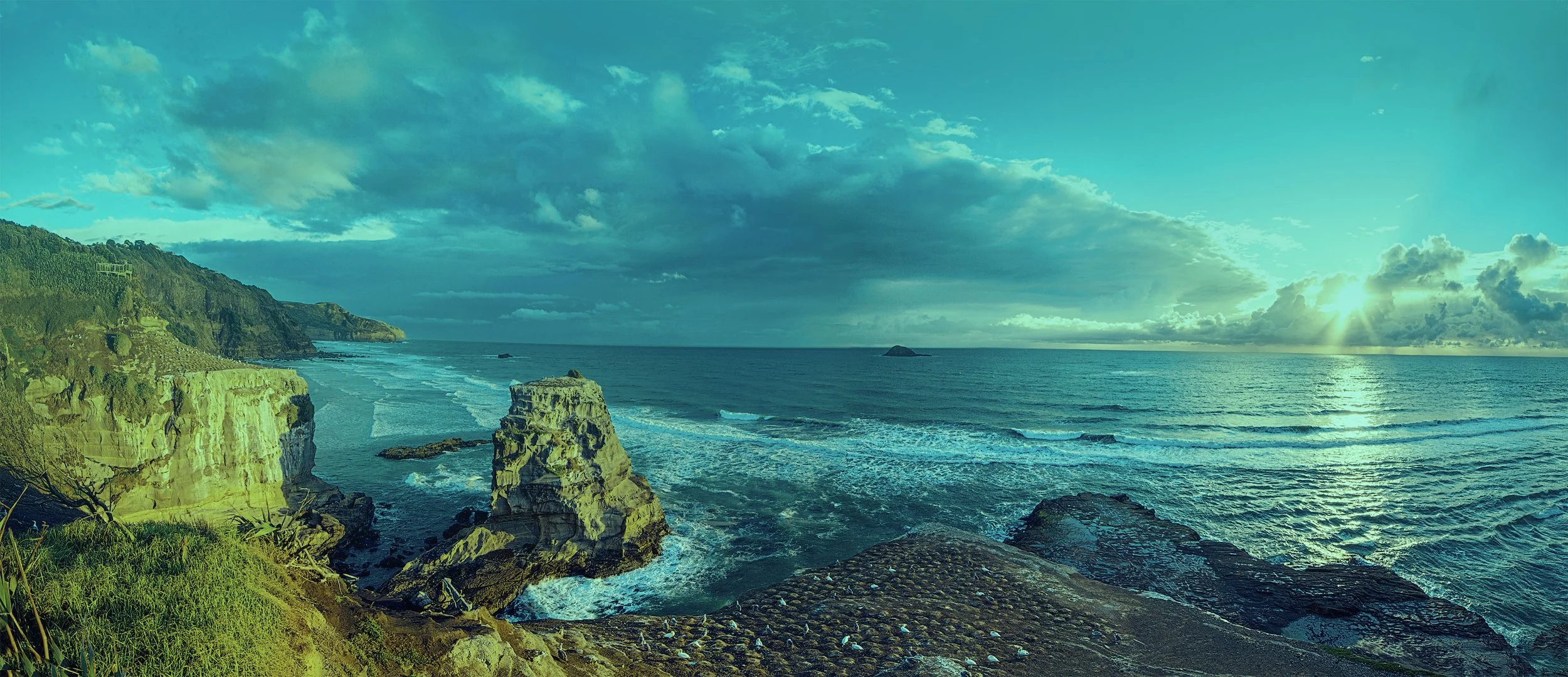 Sunset over the ocean with clouds, rocky cliffs, and green vegetation.