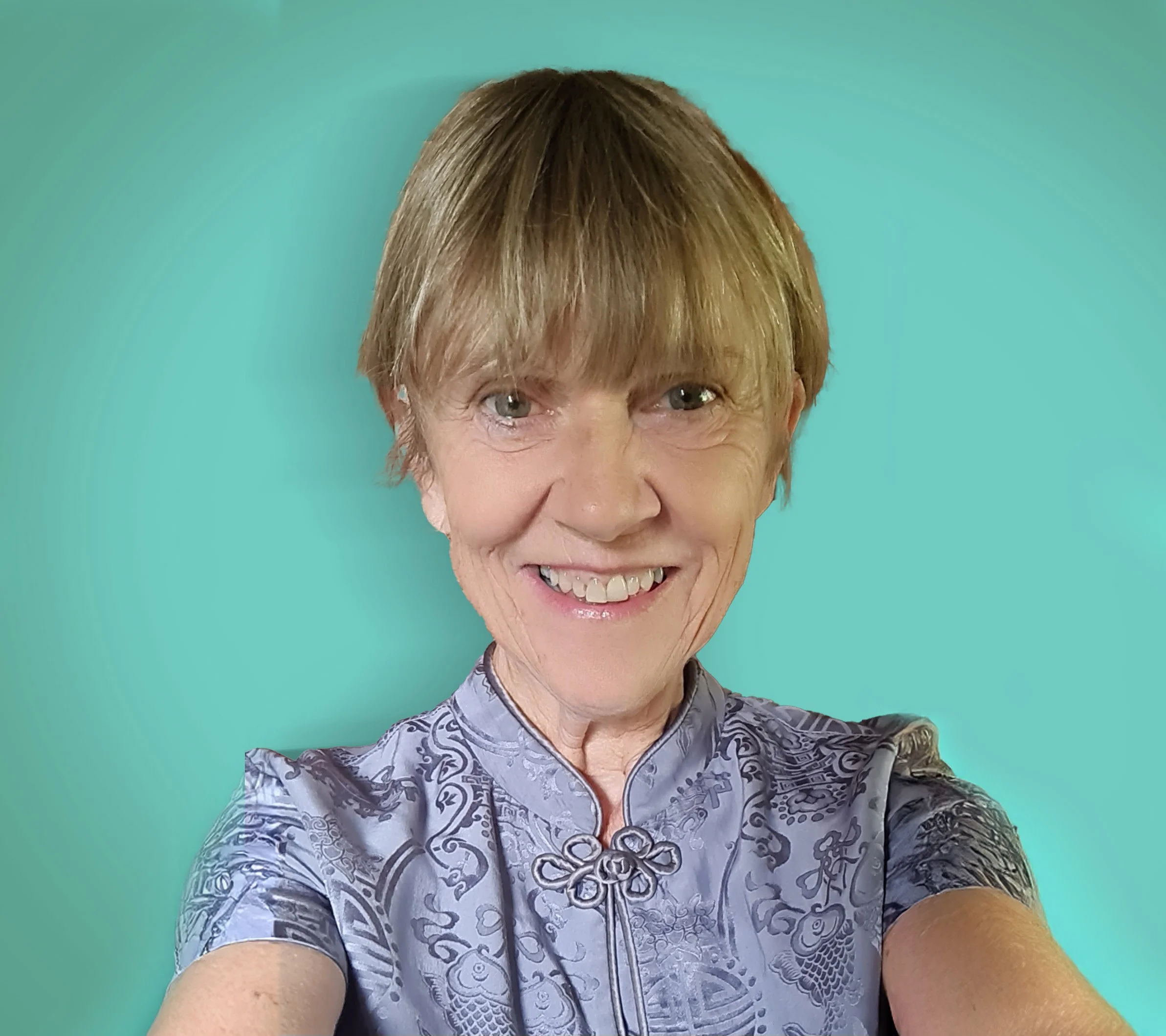 A woman with short blonde hair smiling at the camera, wearing a light purple patterned top, against a teal background.