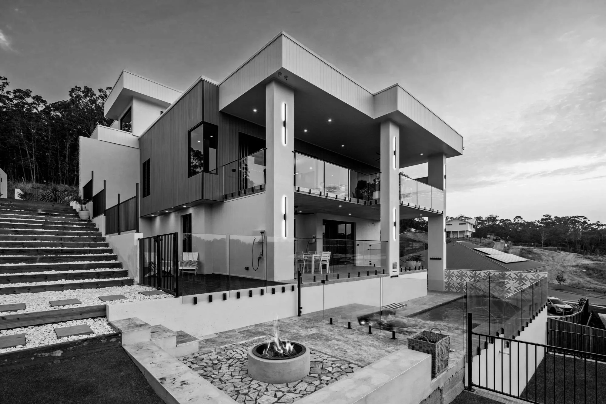 Modern multilevel house with glass railings and exterior lighting, outdoor fire pit, and surrounding landscape with stairs and fencing in black and white.