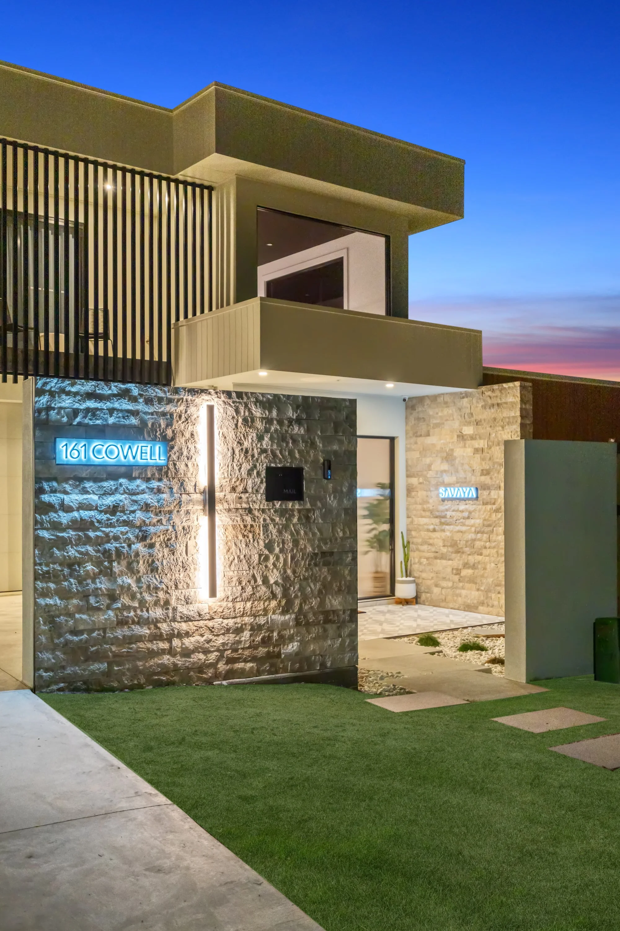 Modern two-story house with stone and concrete exterior, illuminated blue house numbers '161 Cowell', and a small front yard with artificial grass during dusk.