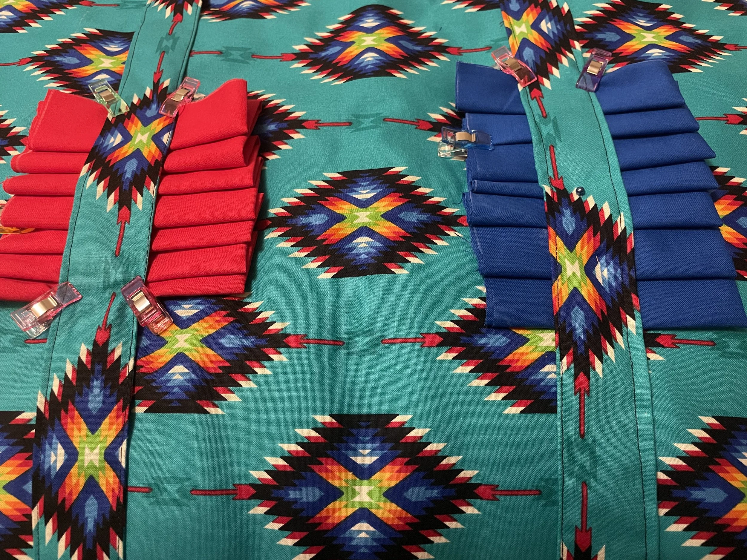Colorful geometric tapestry with two neatly folded stacks of fabric, one red and one blue, secured with fabric strips and small clips, on a vibrant patterned background.