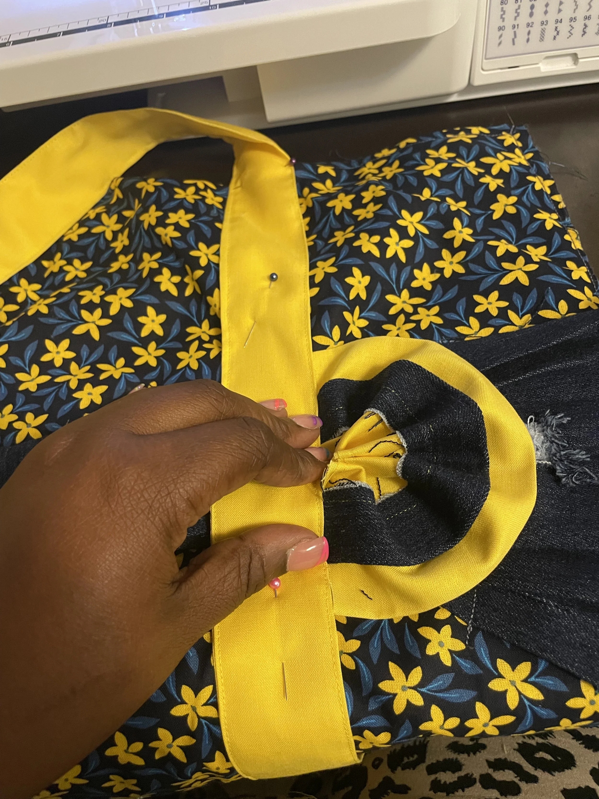 Close-up of a person sewing a piece of fabric with a yellow border and a floral pattern, with the person's hand visible and a sewing machine in the background.