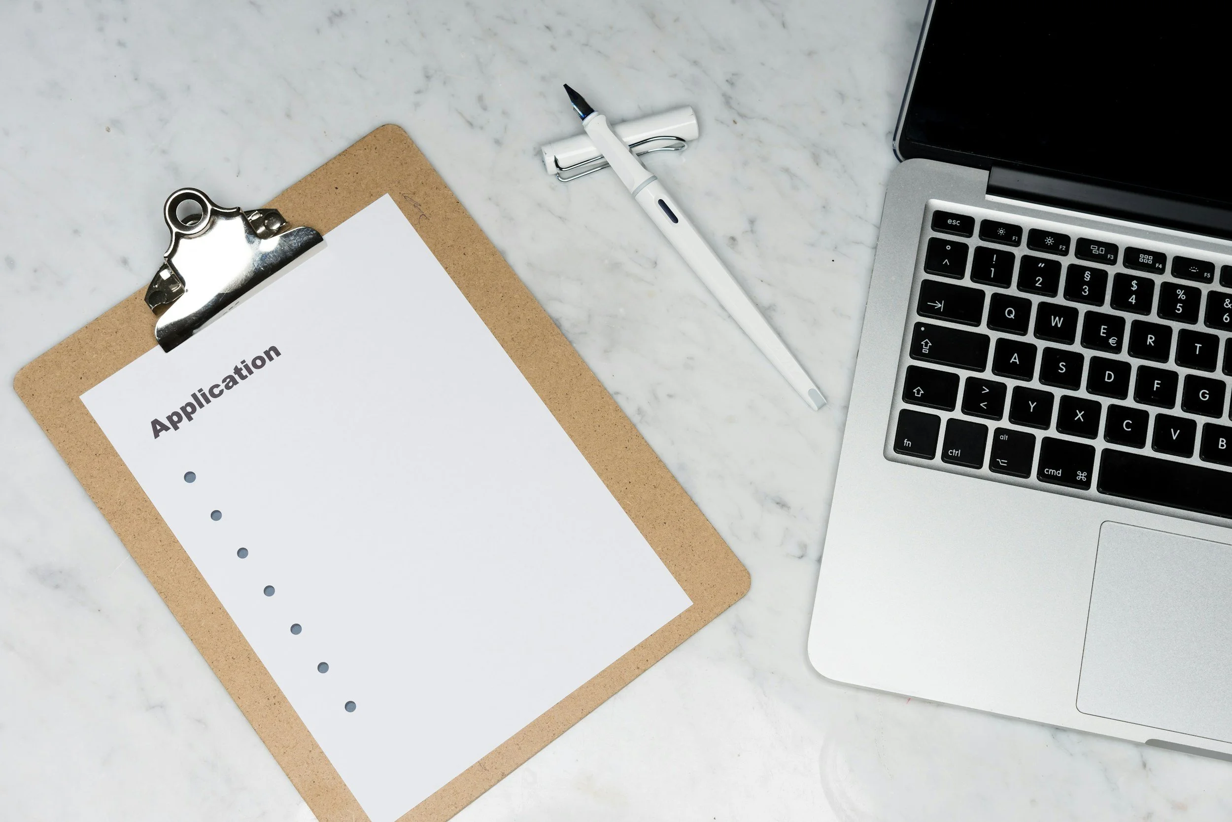 Clipboard with a sheet of paper labeled 'Application,' a white pen, a laptop, and a silver binder clip on a white marble surface.