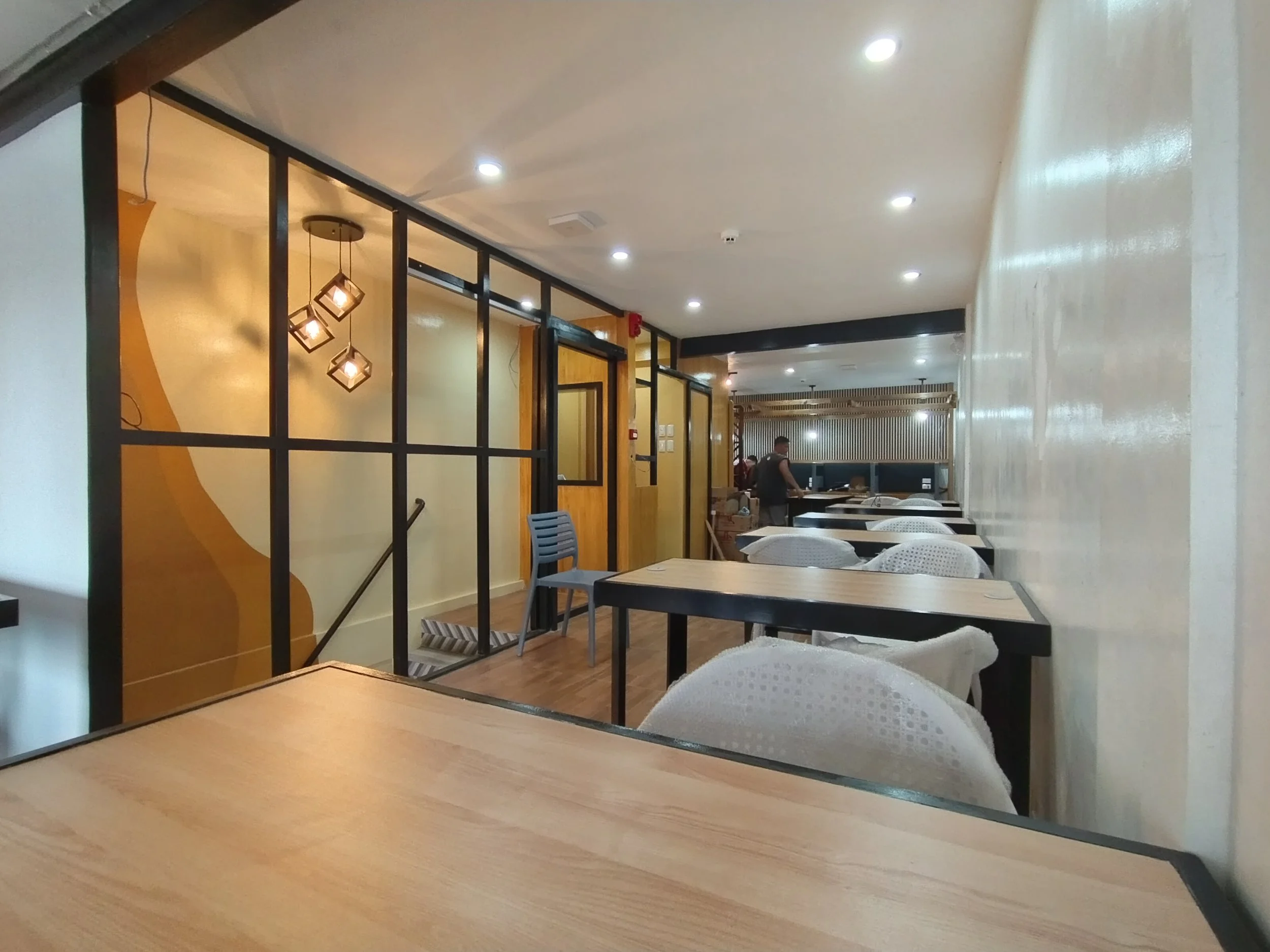 Interior view of a modern restaurant with wooden tables, white chairs, and decorative pendant lights, featuring a partial partition with black frames and a person working in the background.
