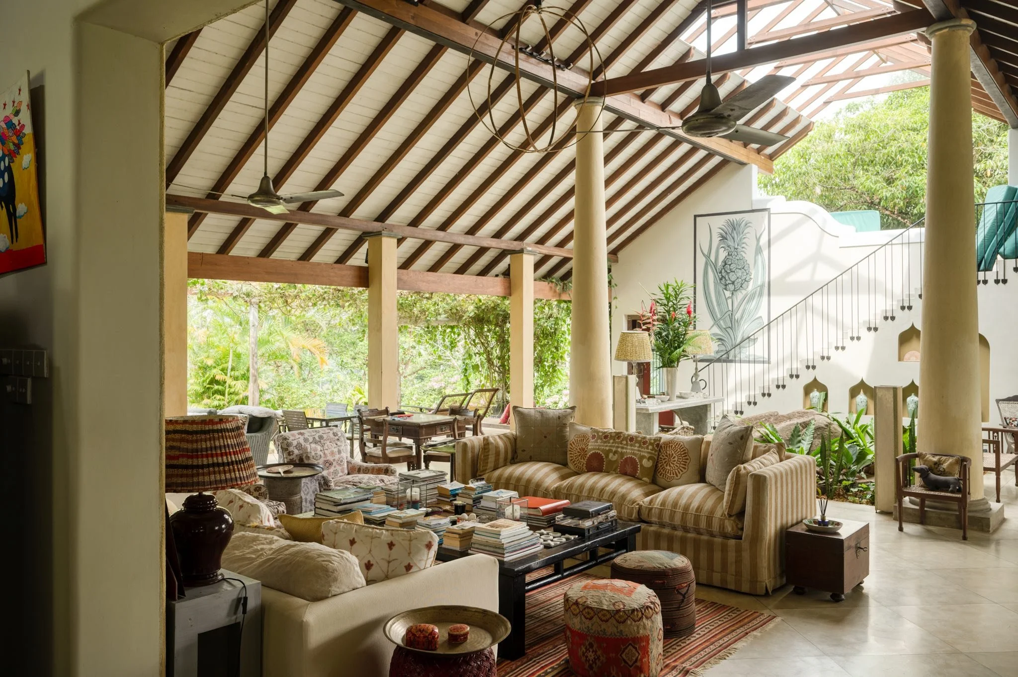 A spacious living room with open walls and a high, sloped wooden ceiling. The room is filled with comfortable furniture including sofas, armchairs, and a coffee table cluttered with books. There are bookshelves, lamps, plants, and artwork, with large windows and an open patio area visible in the background, allowing natural light to fill the room.