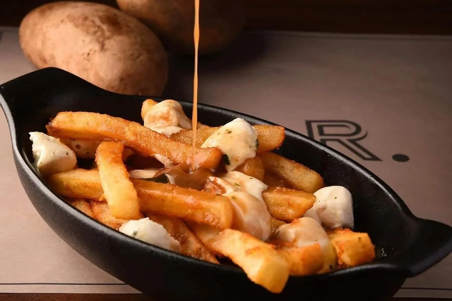 Poutine - French fries topped with cheese curds in a black dish, with hot gravy being poured over them from a glass gravy boat, and potatoes in the background.