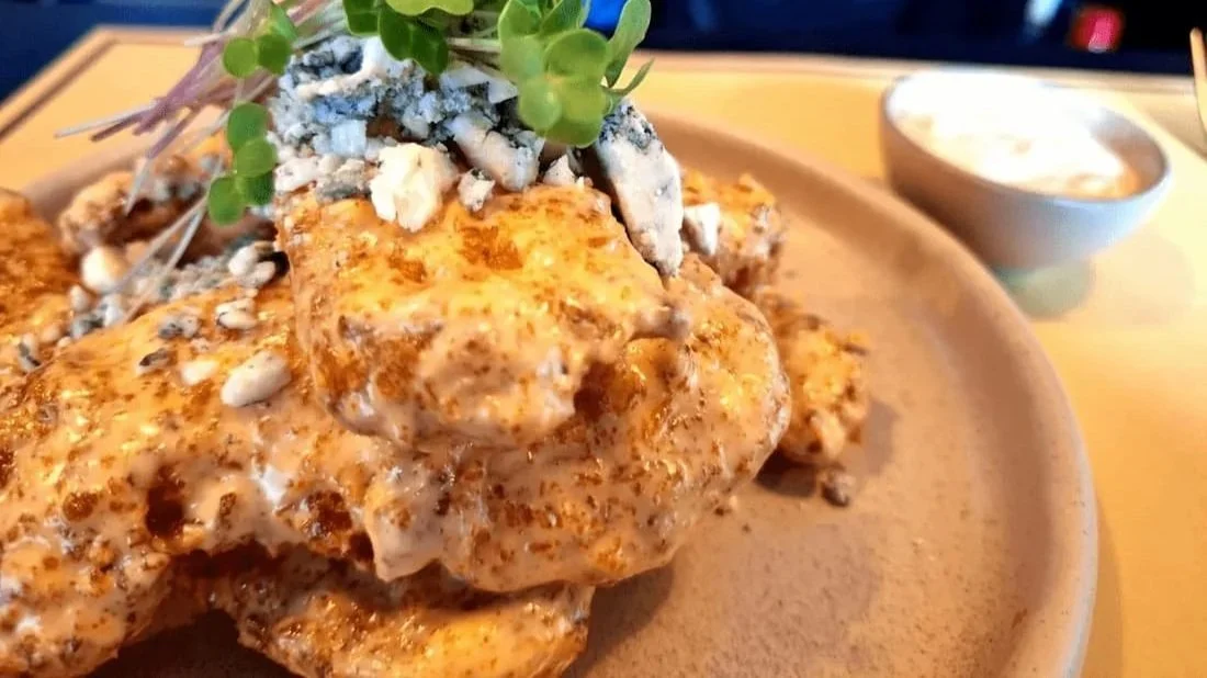 Close-up of fried chicken topped with crumbled blue cheese, microgreens, and seasoning, served with a side of dipping sauce.