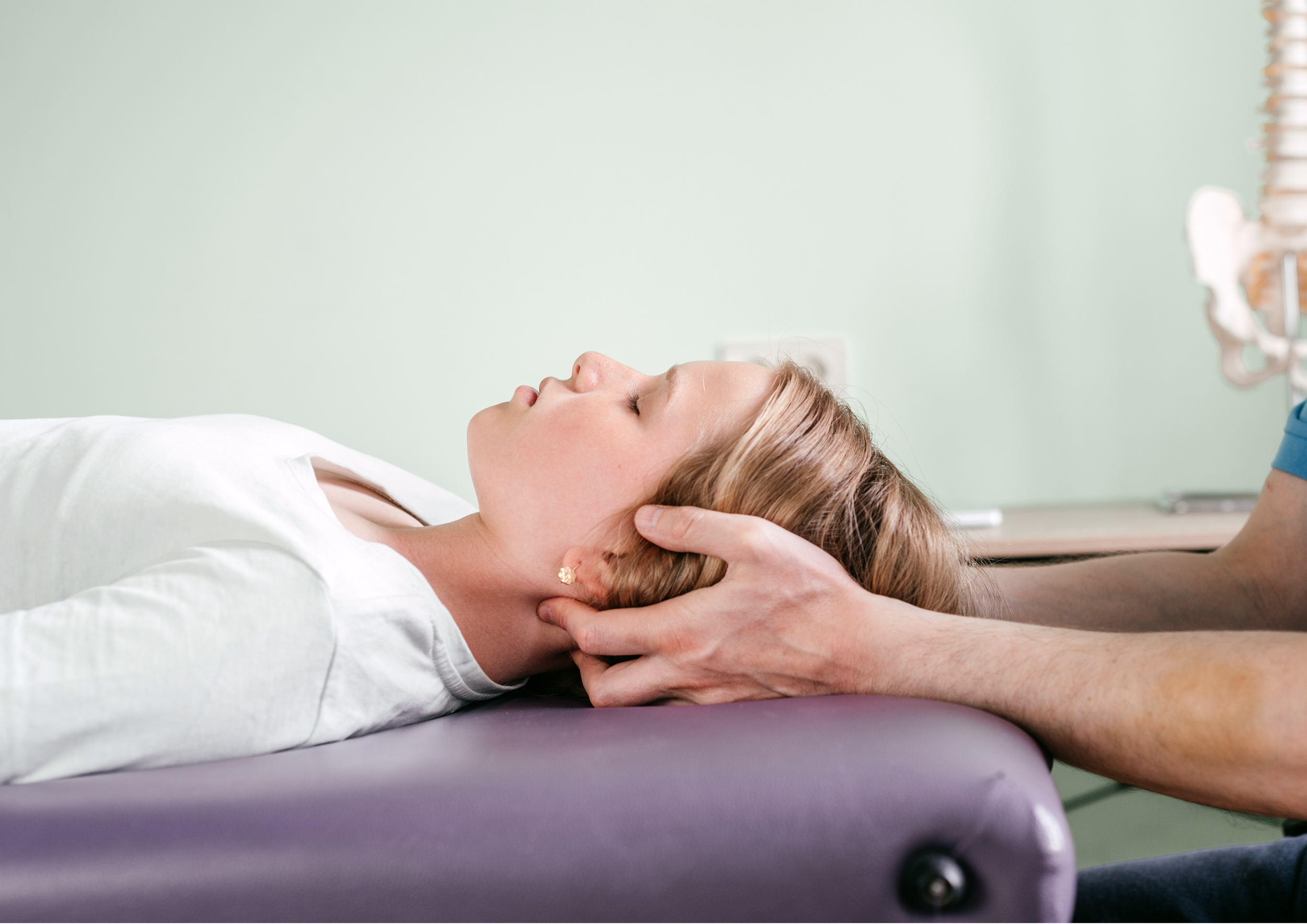 A woman lying on her back on a treatment table with her eyes closed, receiving a neck examination or therapy from a healthcare professional.